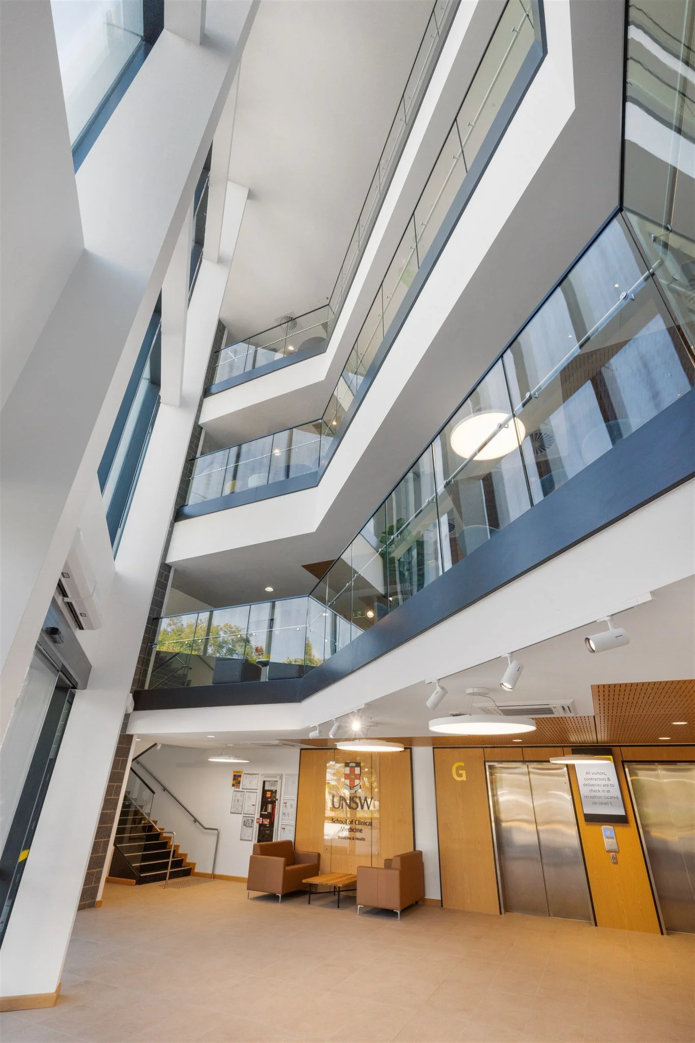 Interior view of a modern multi-floor building with glass balconies, staircase, and seating area. Signage for UNSW School of Clinical Medicine is visible on a wooden wall.