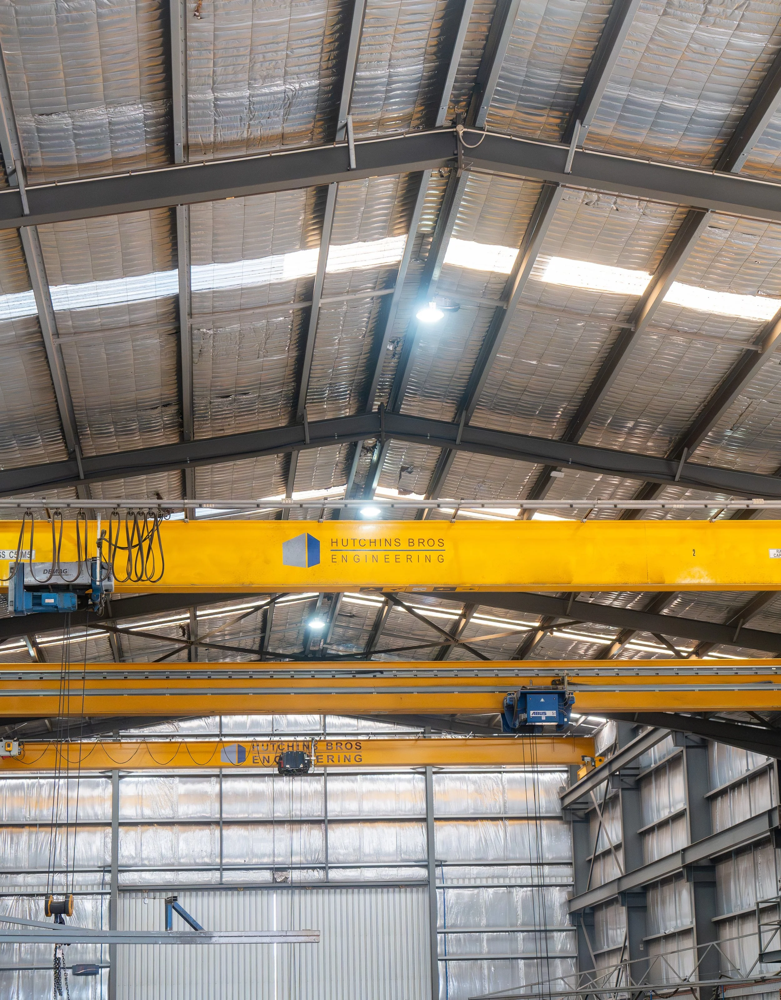 Interior view of a large industrial building with high metal ceiling, steel beams, and a yellow overhead crane labeled 'Hutchins Bros Engineering'.