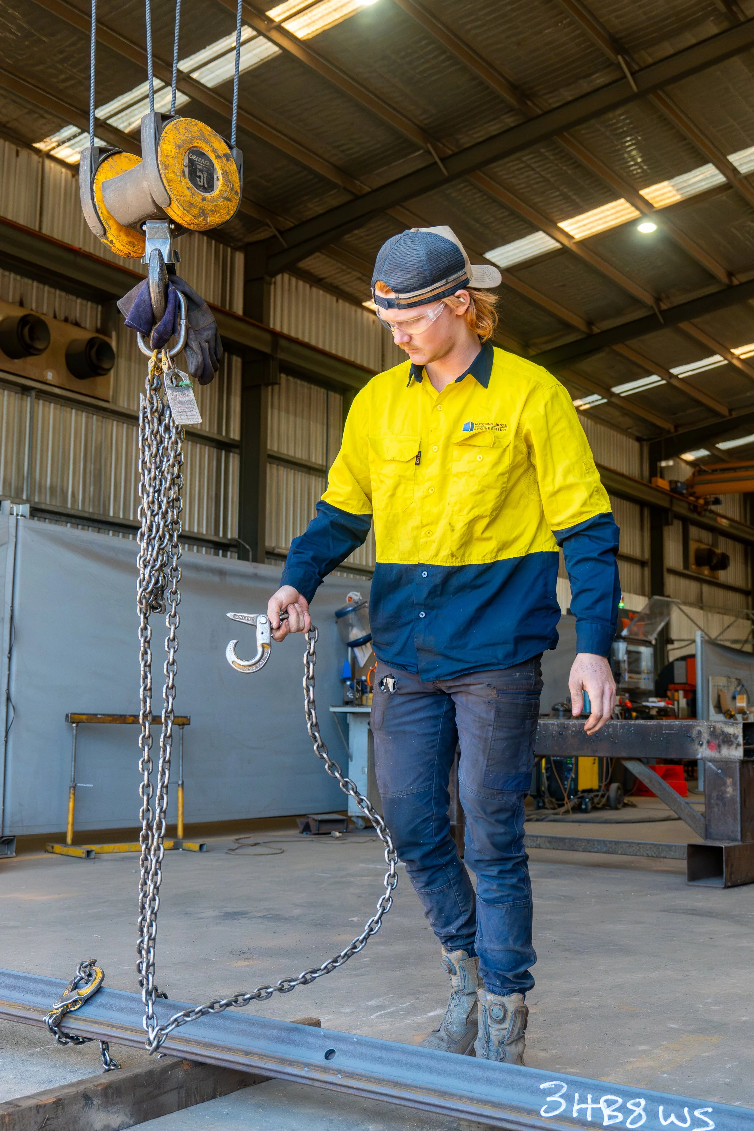 A worker in safety gear operating an overhead crane using the hook and chain to pick up a steel assembly inside Hutchins Bros workshop.