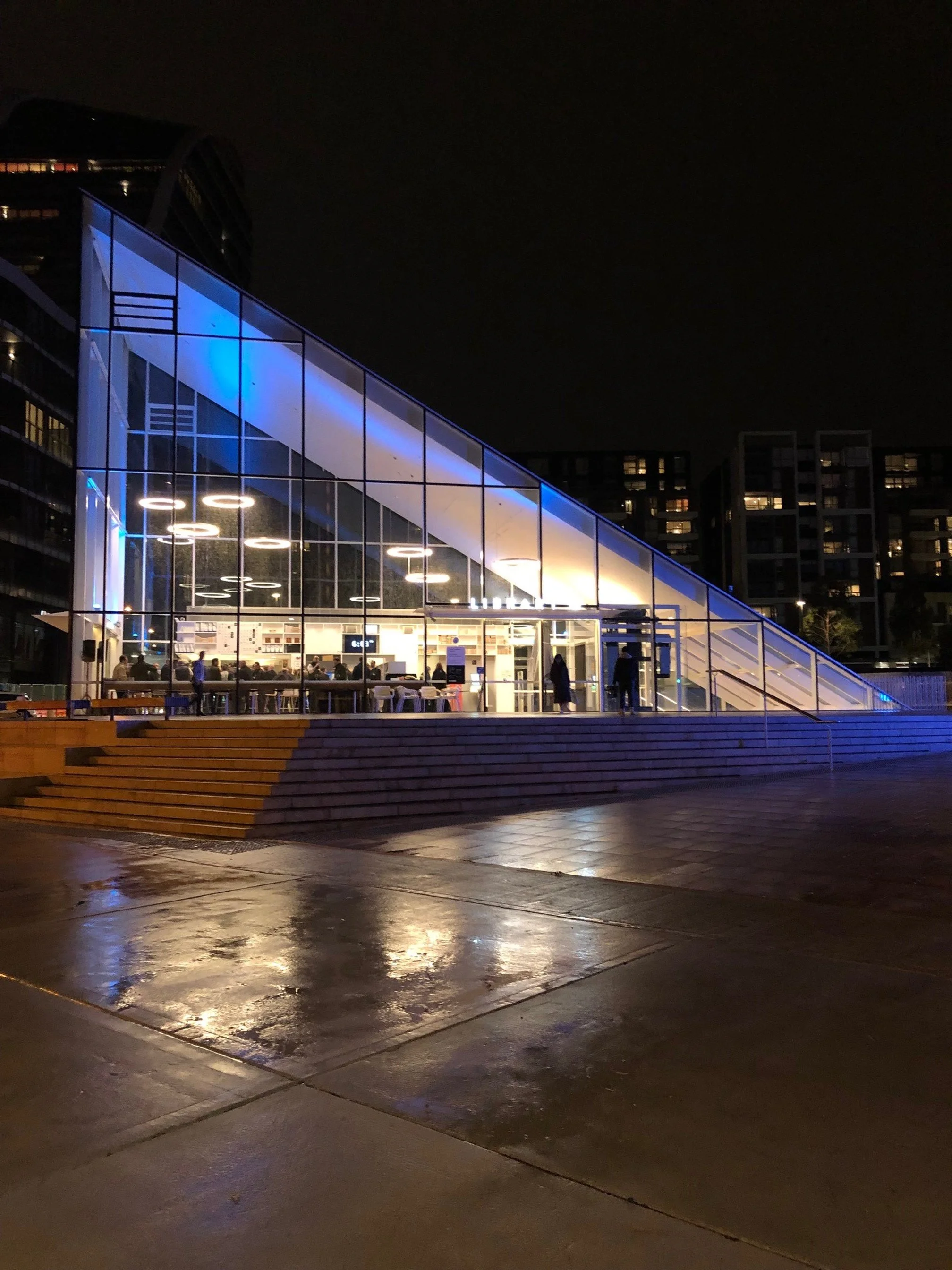 Night view of a modern glass building with interior lighting, showing people sitting inside, with wet pavement reflecting lights in the foreground.