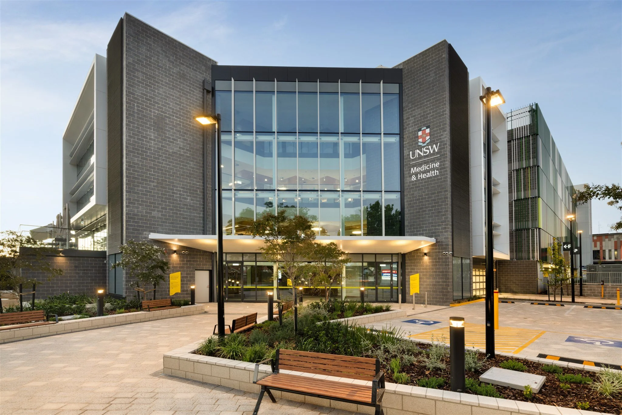 Modern medical building with glass windows and the UNSW Medicine & Health sign, surrounded by greenery, benches, and lamp posts, during early evening.
