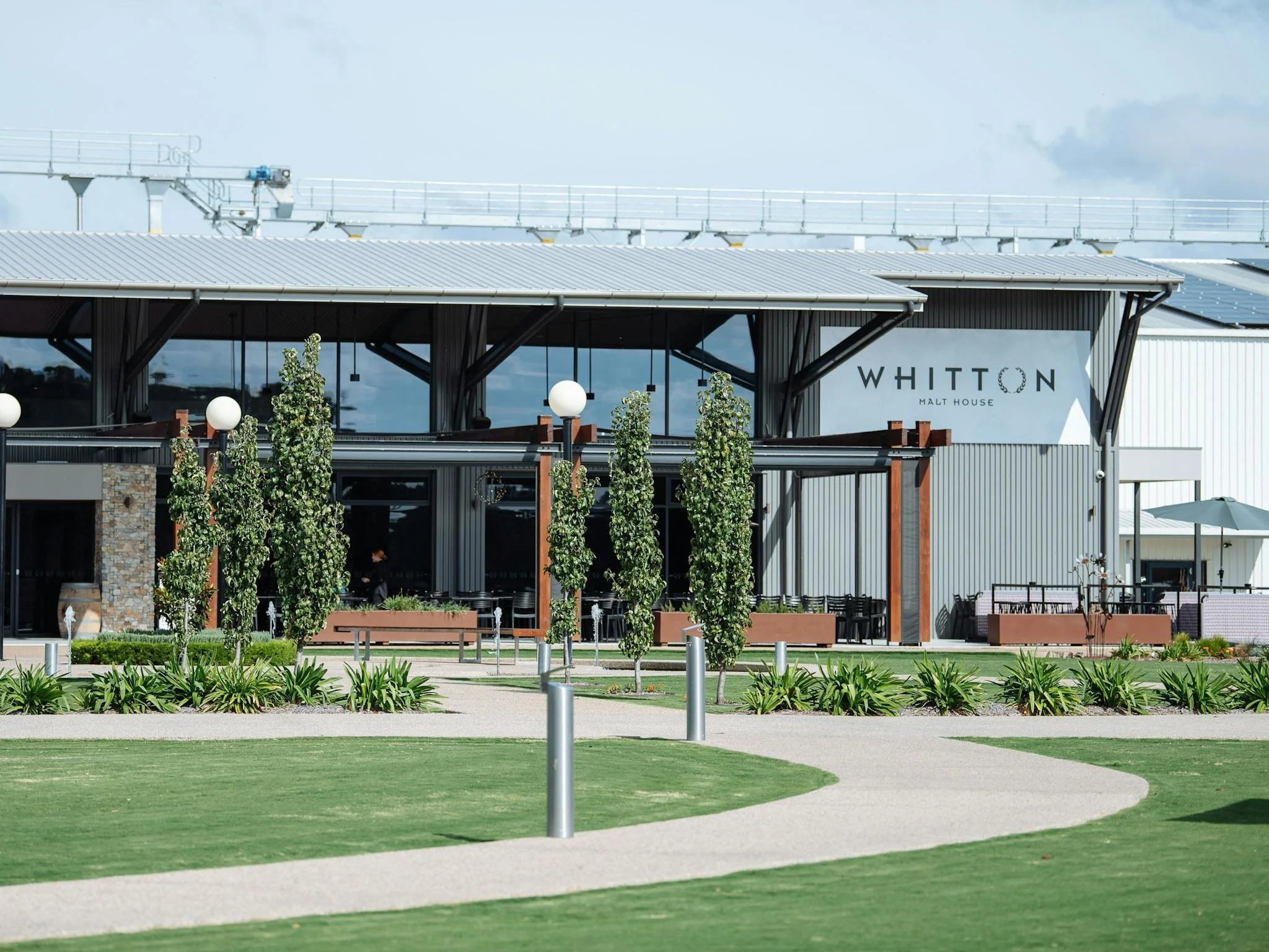 A modern building with a sign that reads 'Whiton Malt House,' surrounded by a landscaped area with rowed trees, lampposts, and a grassy area with pathways.