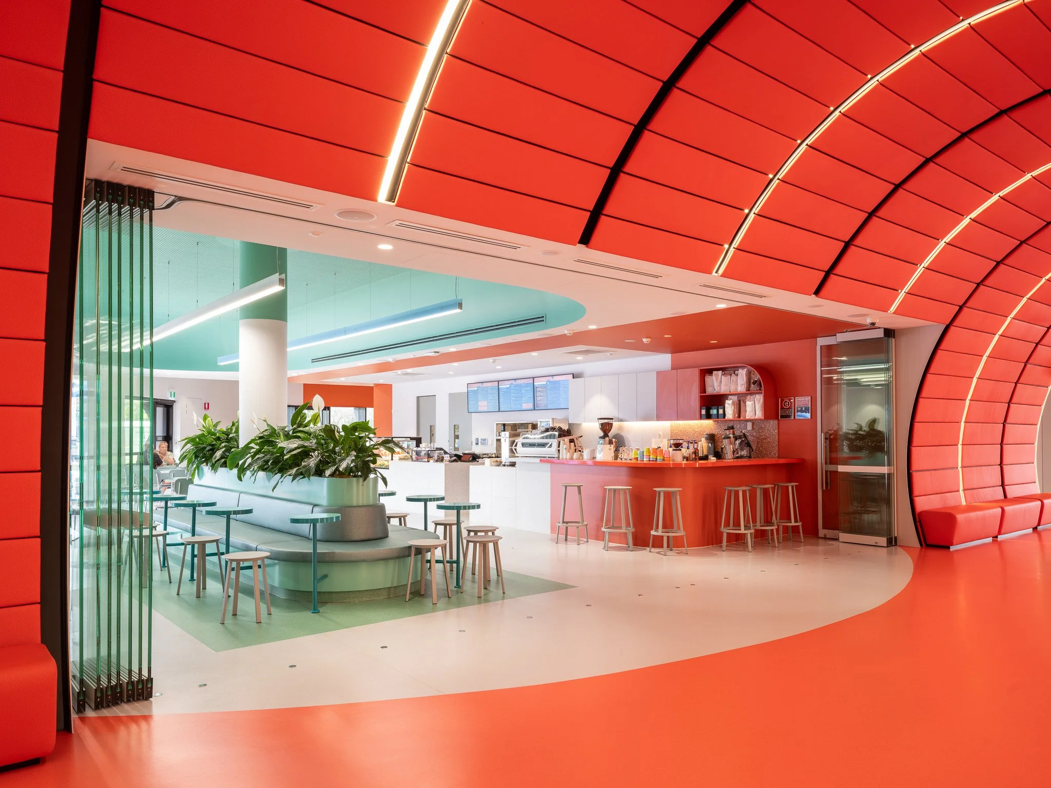 Modern interior of a cafe or lounge with red, orange, and light teal color accents, seating areas, and a counter for ordering food and drinks.