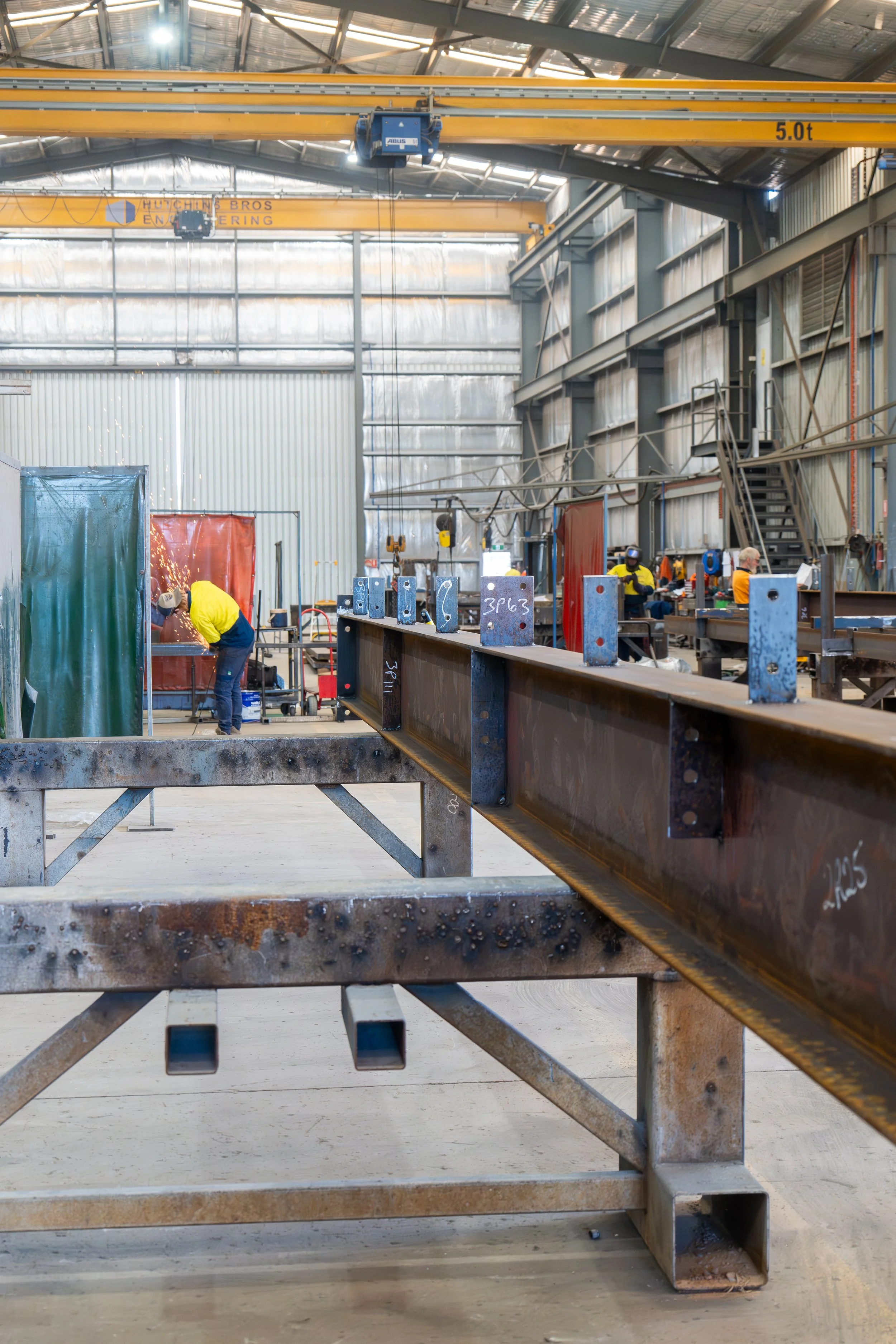 View of an industrial workshop with workers welding metal beams, large yellow overhead cranes, and a metal staircase.