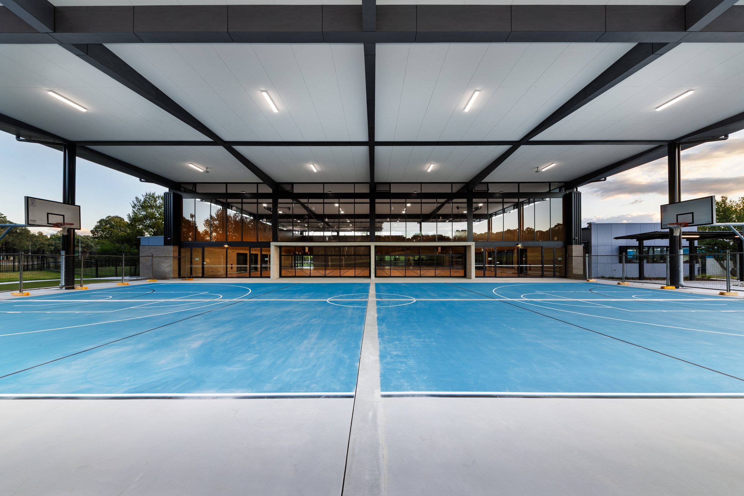 Outdoor basketball court with a bright blue surface, white boundary lines, and two basketball hoops, with a modern glass-fronted building in the background.