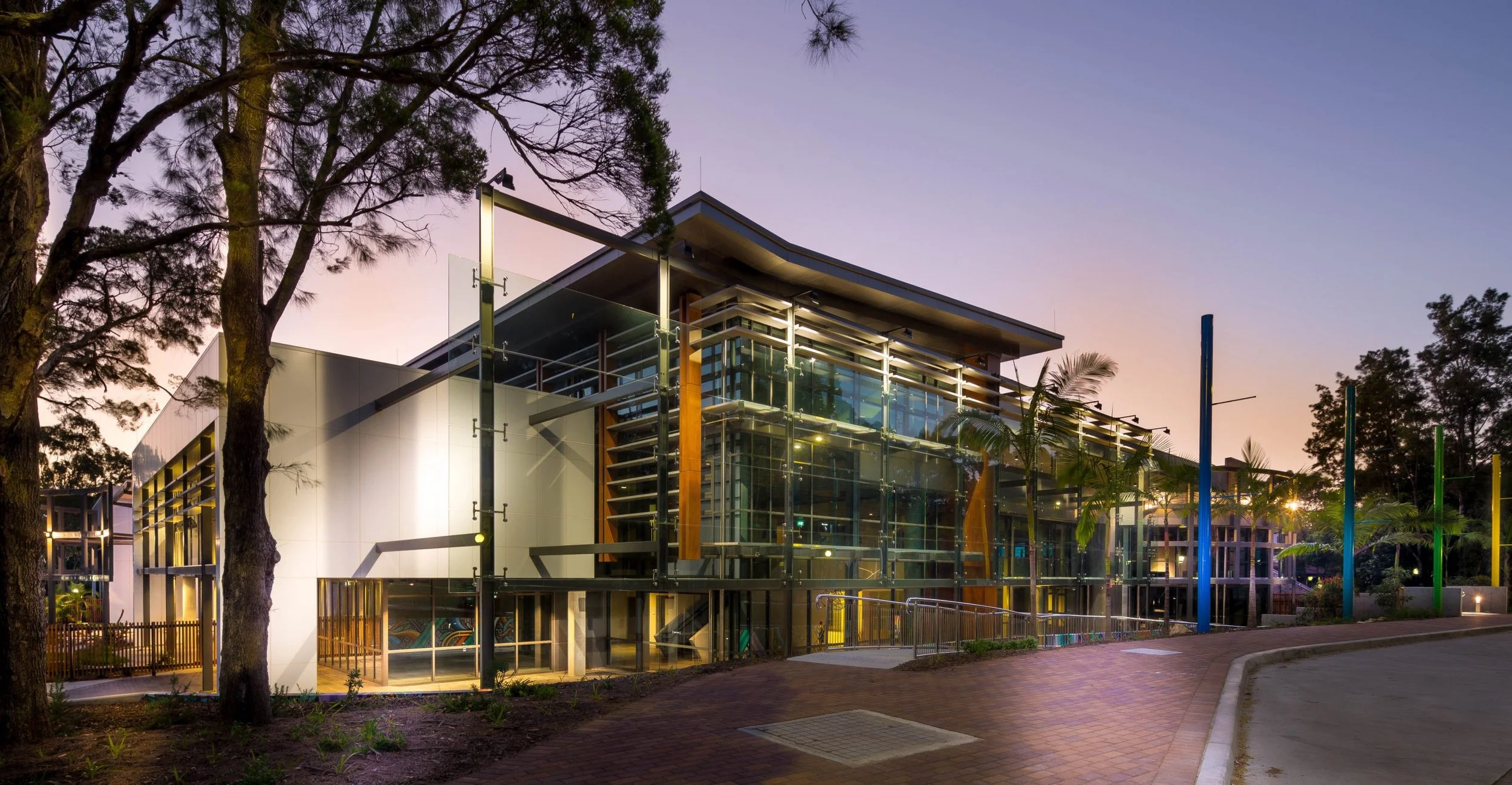 Modern glass building at dusk with trees and colorful vertical poles outside.