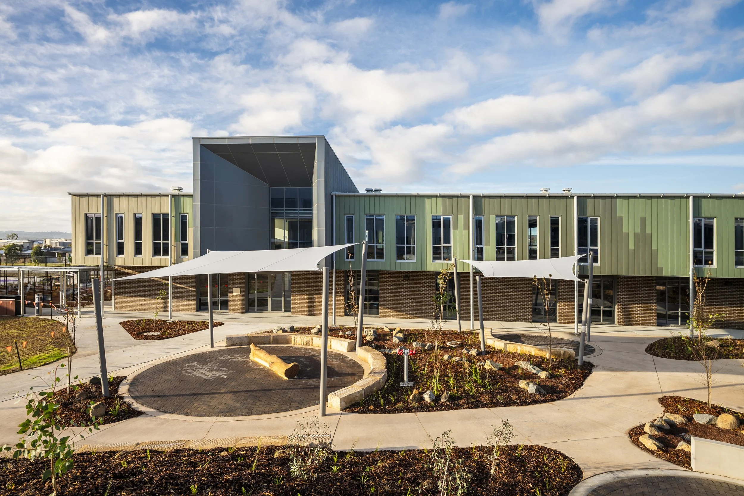 Modern school building with green and beige exterior, large windows, and a courtyard with circular paved walkway, garden beds, and shade sails against a partly cloudy sky.