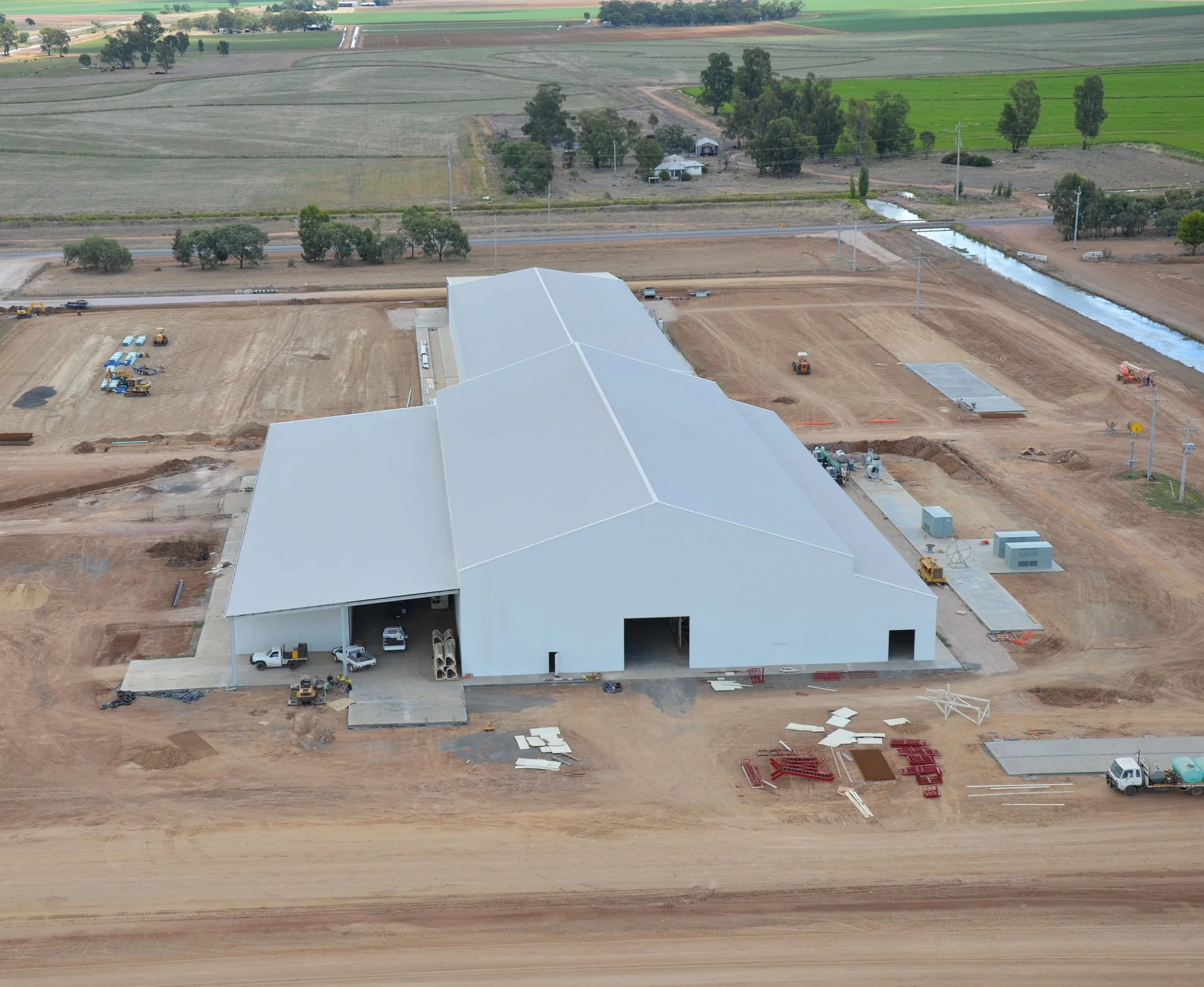 Construction site with a large building under construction, surrounded by dirt and construction equipment, with a rural landscape of fields, trees, and a water channel in the background.