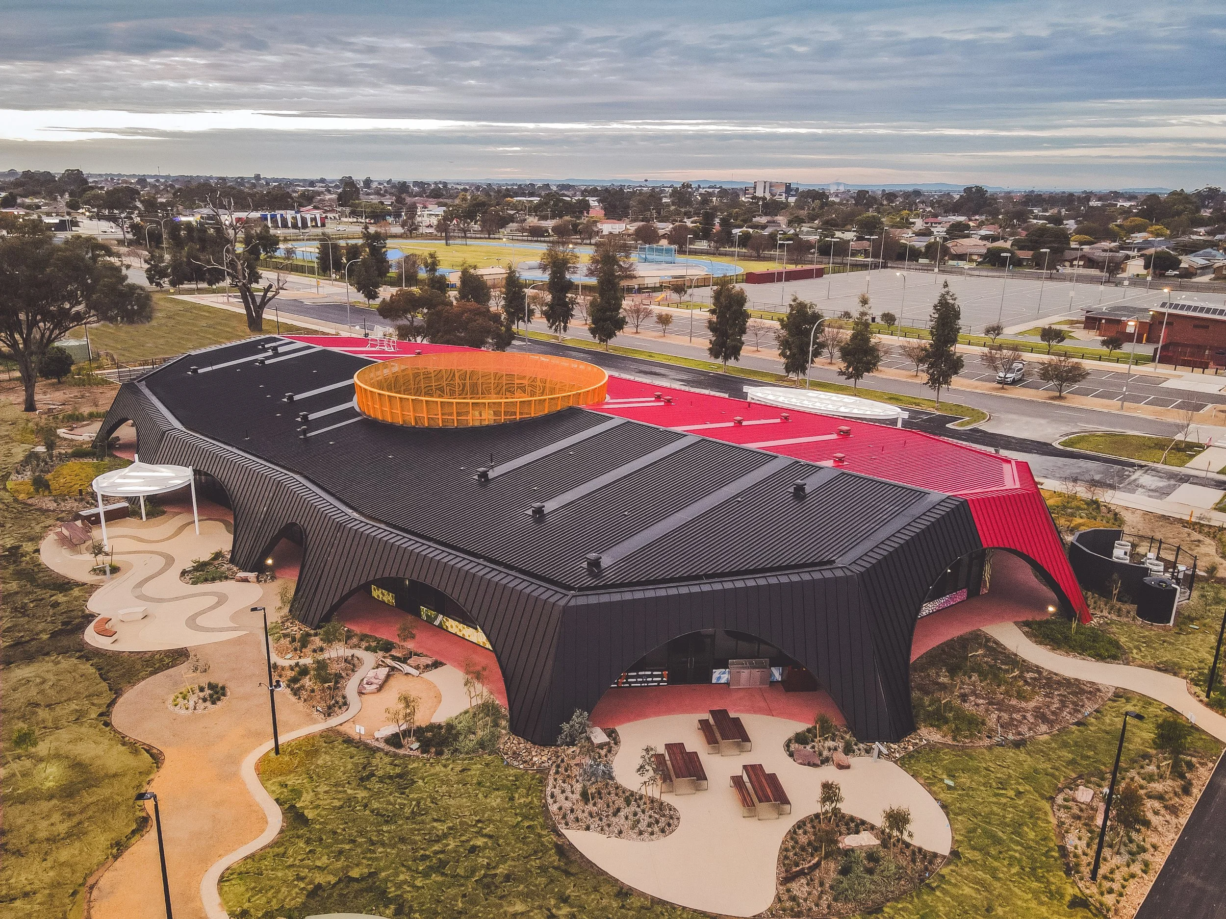 Aerial view of a park with a black curved roof building, orange circular structure, walking paths, trees, open fields, and a parking lot.