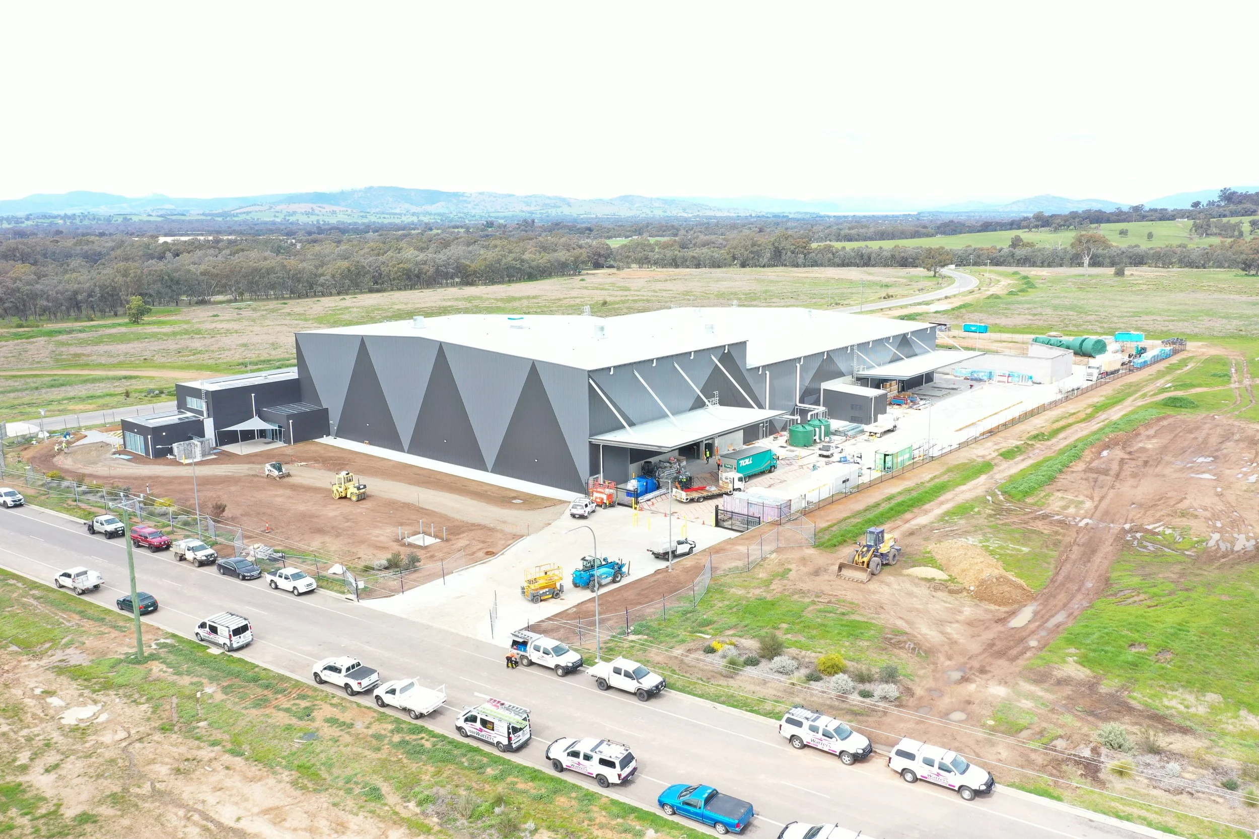 An aerial view of an industrial building with a white roof surrounded by a rural landscape with open fields, trees, and a winding road. Several parked vehicles and construction equipment are visible around the building.