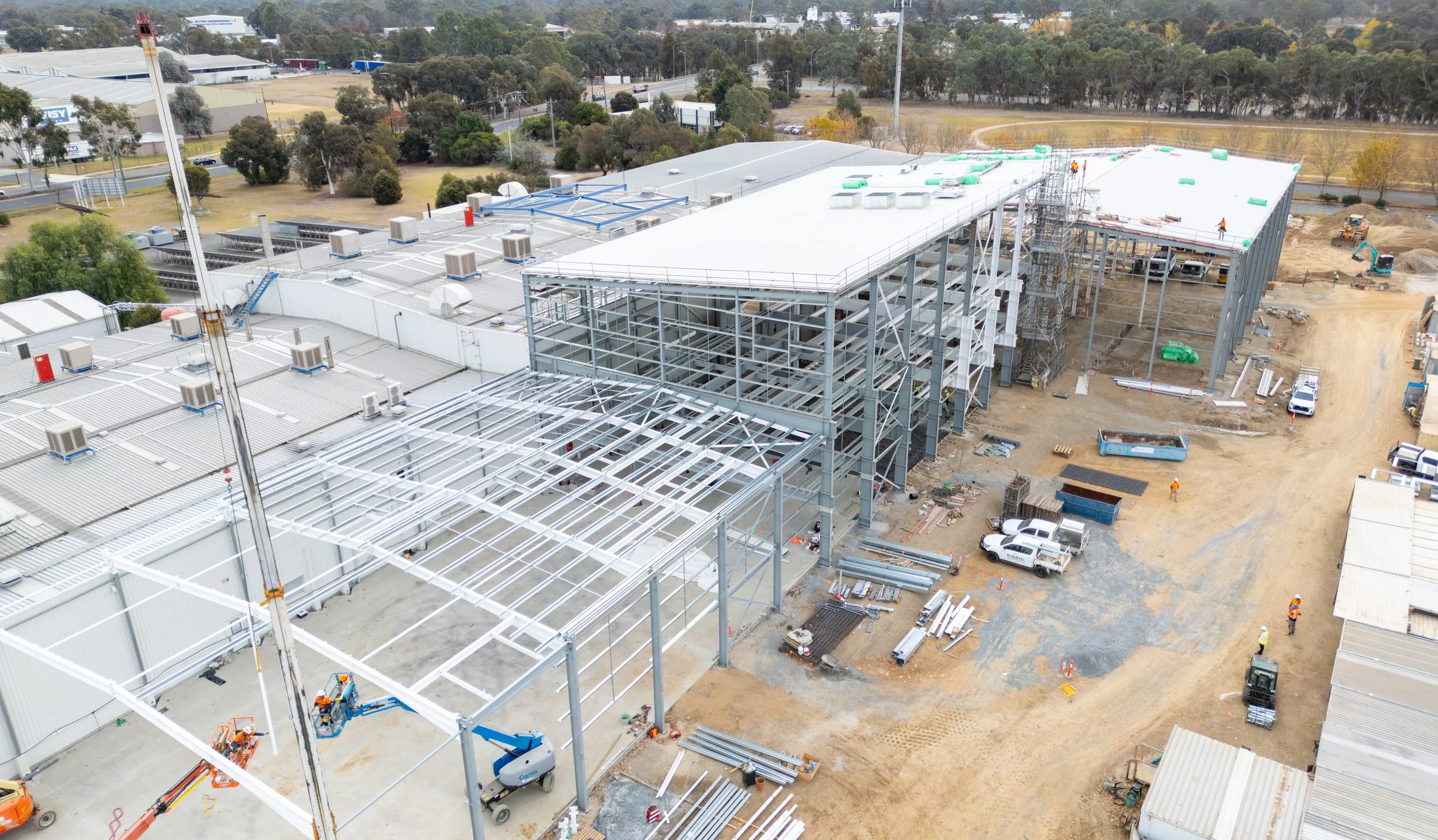 Construction site with a large building under construction, steel framework, workers, and construction vehicles, surrounded by an open area with trees and roadways in the background.