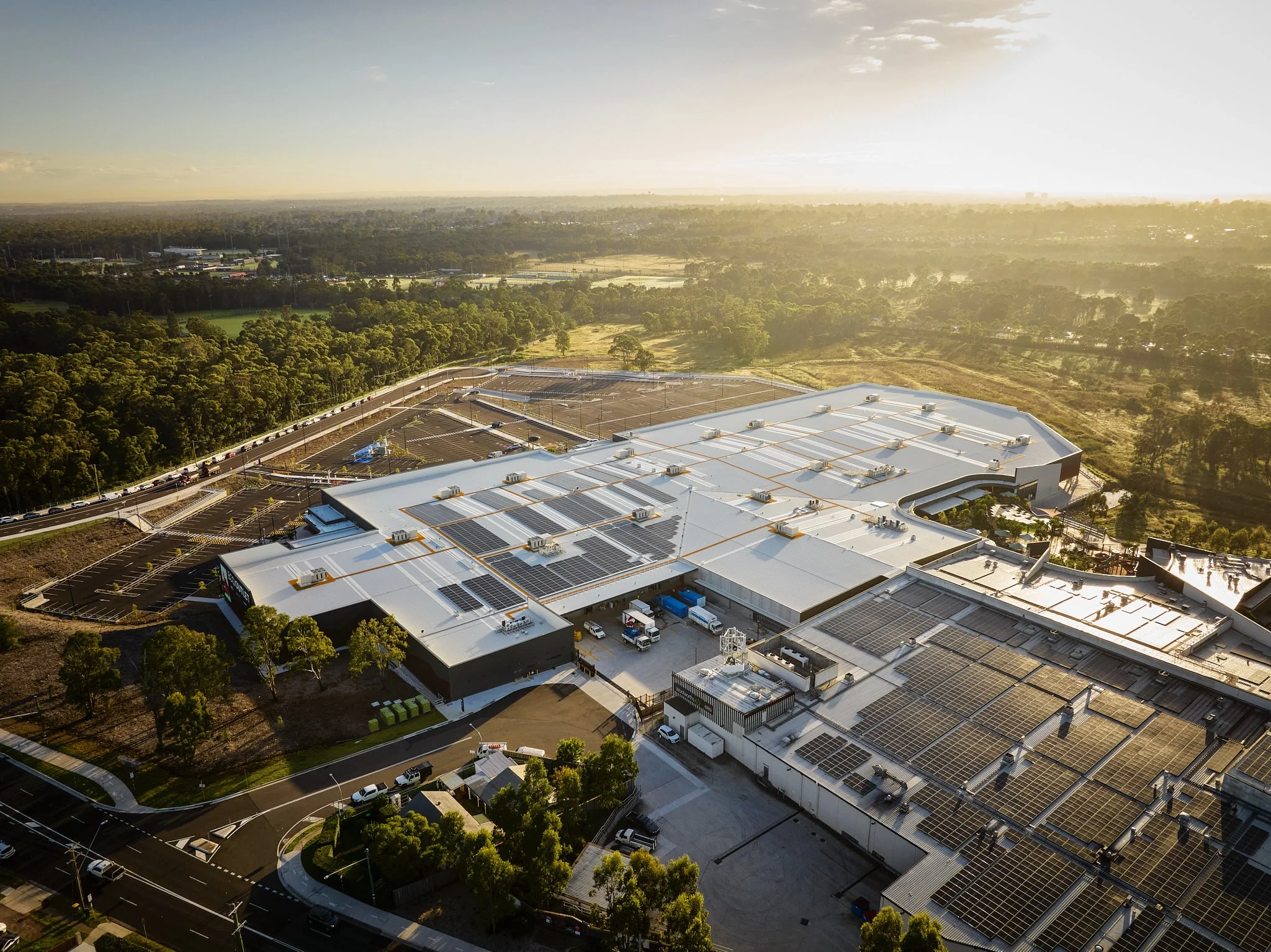 Aerial view of a large commercial building with solar panels on the roof, surrounded by parking lots, roads, and green trees under a clear sky with sunlight.