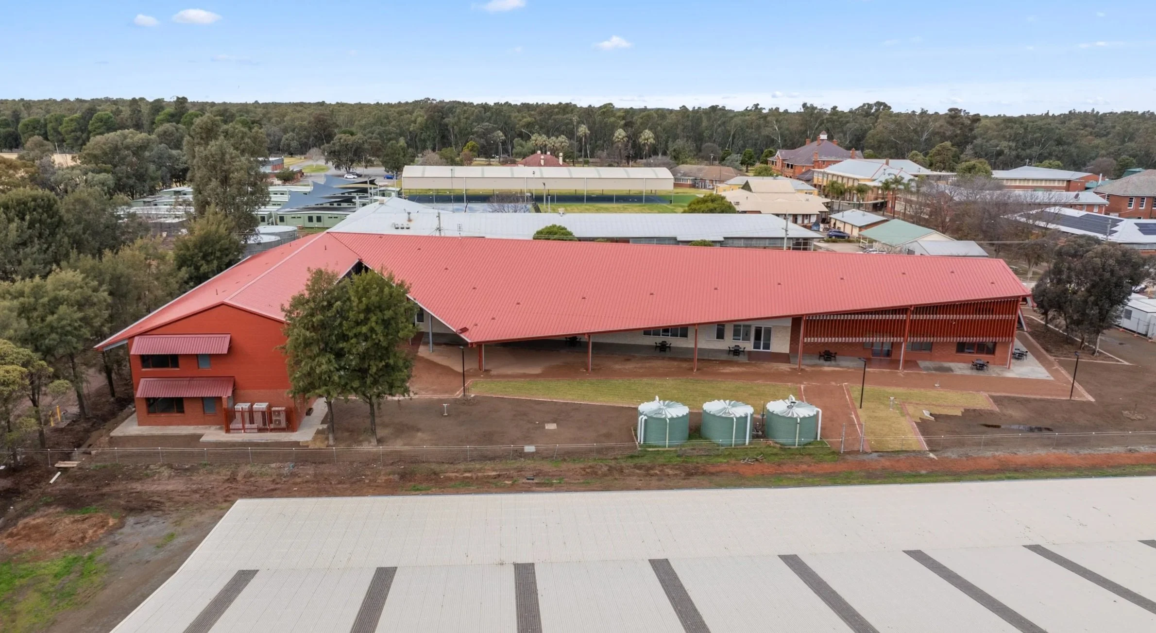 Aerial view of a school campus with various buildings, trees, and a field, with a wooded area in the background and partly cloudy sky.
