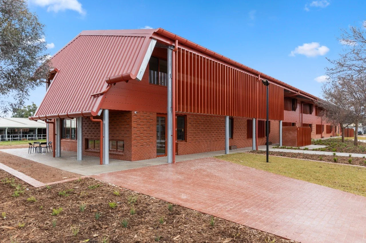 A modern two-story brick building with a red metal roof and vertical slats on the upper level, surrounded by landscaped pathways, grass, and trees under a blue sky.