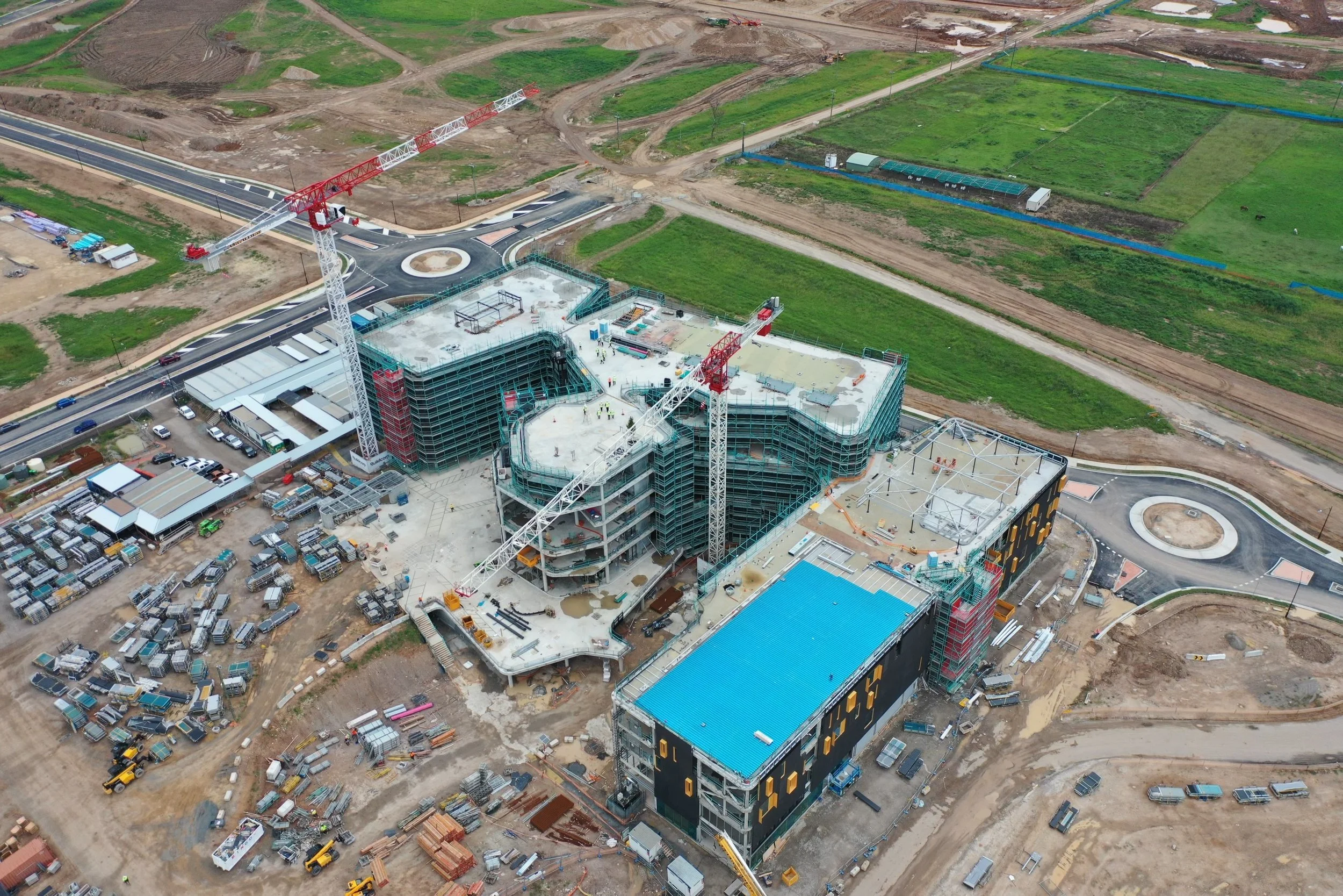 Aerial view of a construction site with buildings under construction, cranes, construction materials, and trucks, surrounded by green fields and dirt roads.