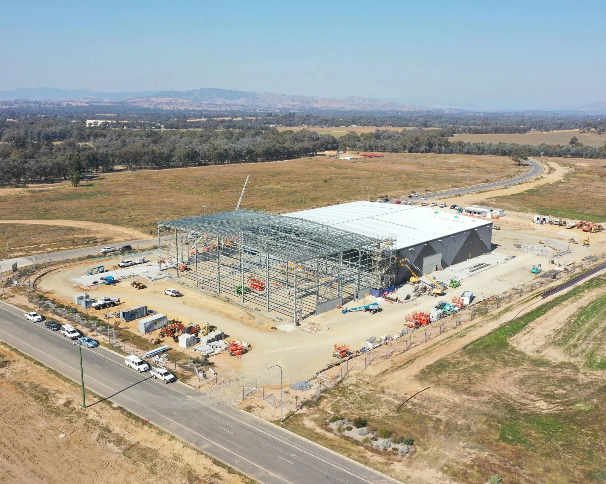 Aerial view of a construction site in a rural area with a steel framework for a building. Surrounding the site are open fields, trees, a curved road, and parked vehicles. In the background, there are hills under a clear blue sky.