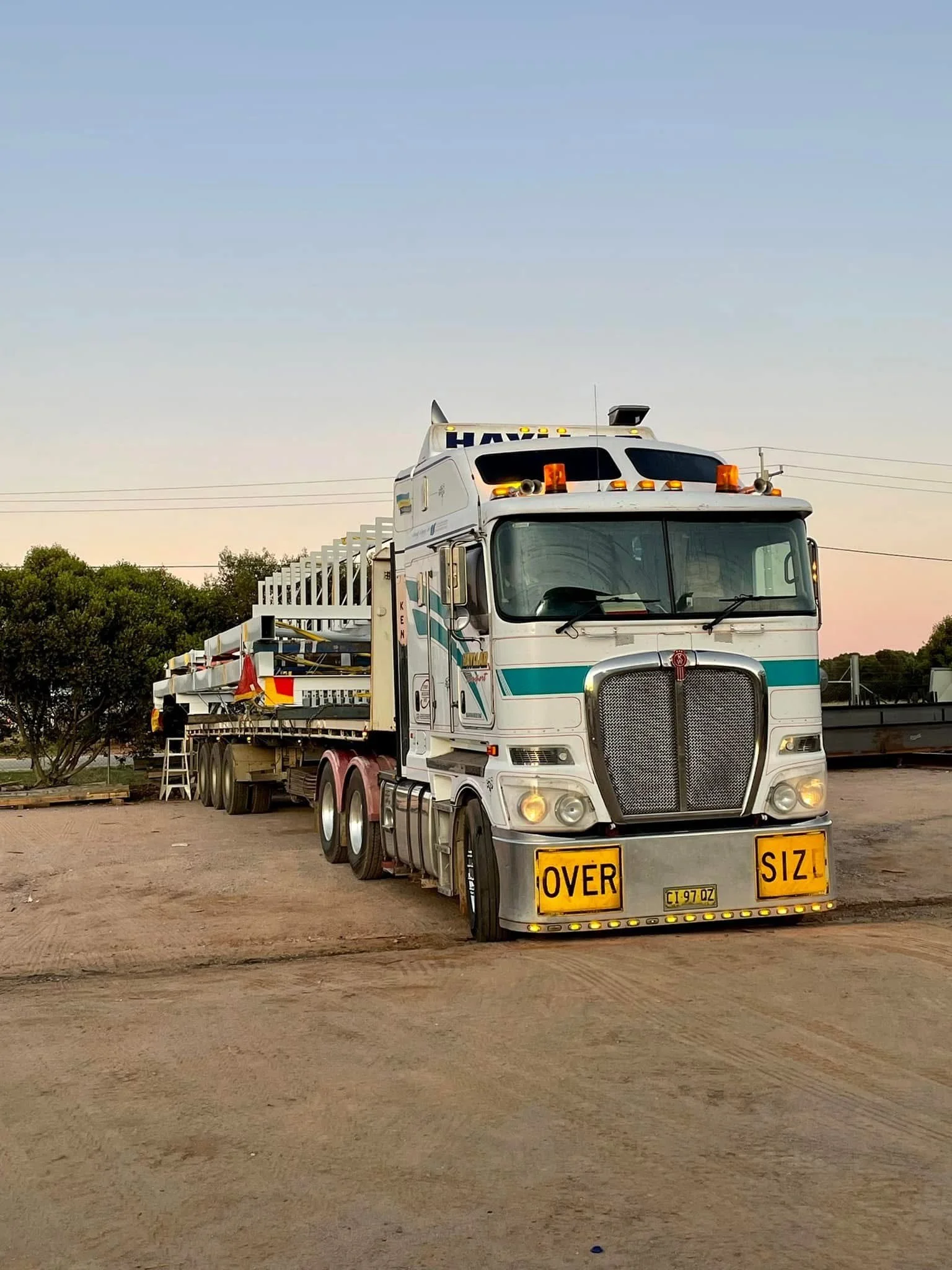 A large white semi-truck with yellow and black signs that say "OVER SIZE" on the front bumper, parked on a dirt lot with trees and power lines in the background during sunset.