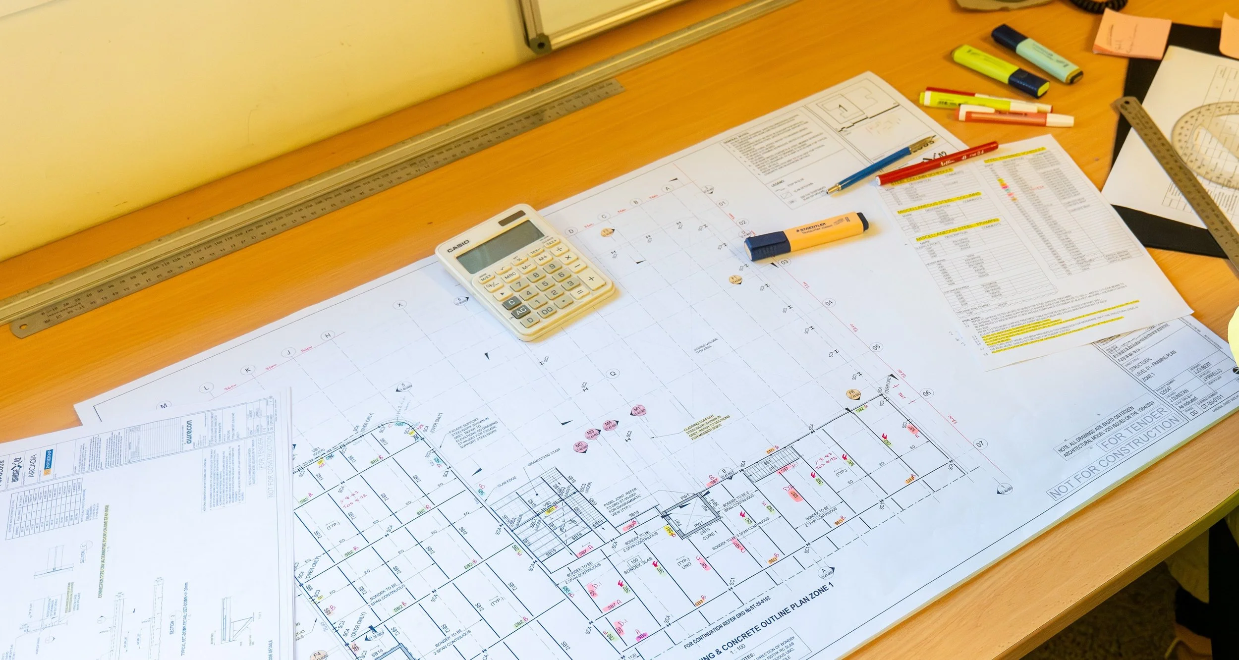 Architectural blueprints and construction plans spread out on a wooden table with various highlighters, a calculator, and pens for reference.