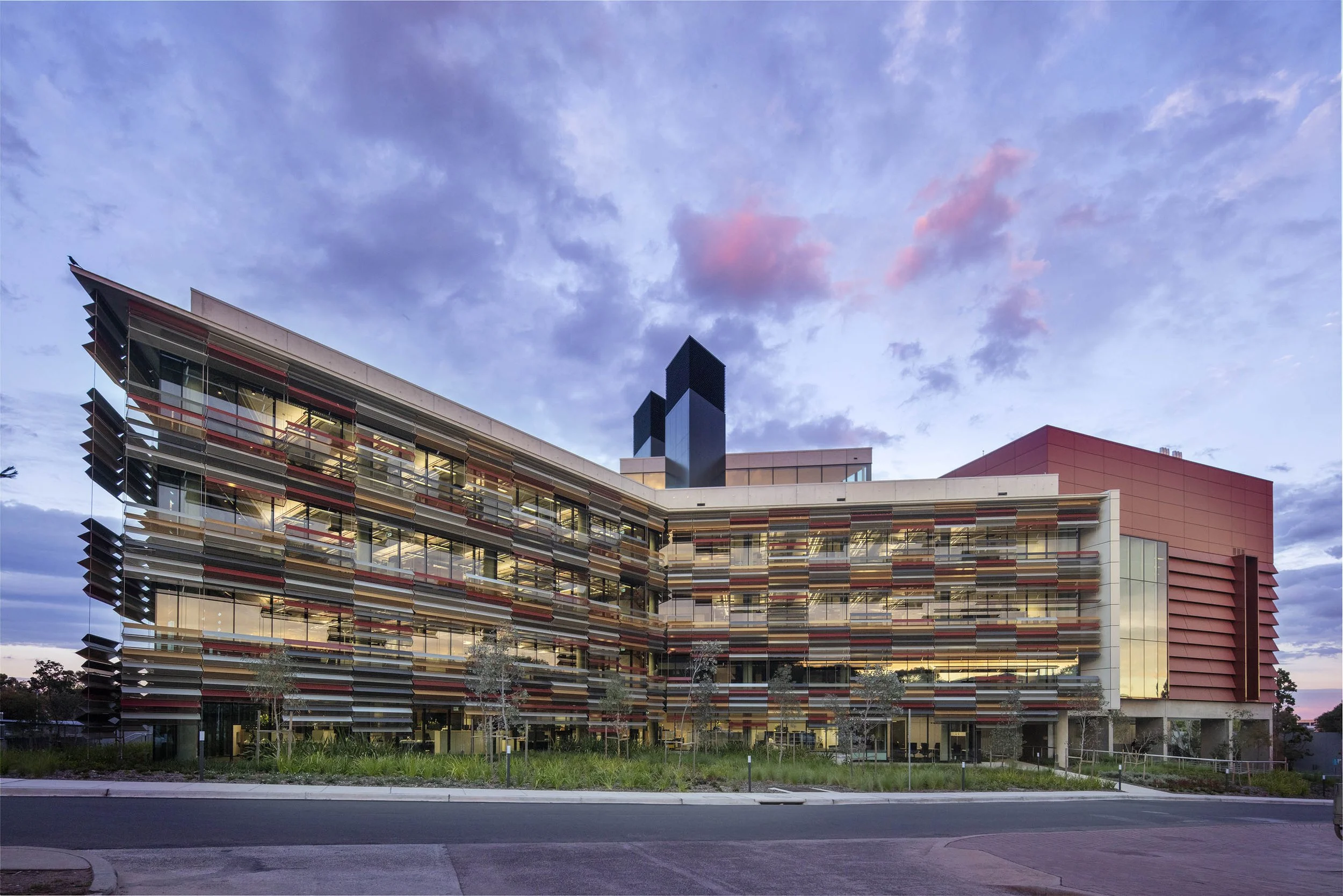 Modern multi-story building with colorful horizontal slats, large glass windows, and a rooftop structure, set against a evening sky with pink and purple clouds.