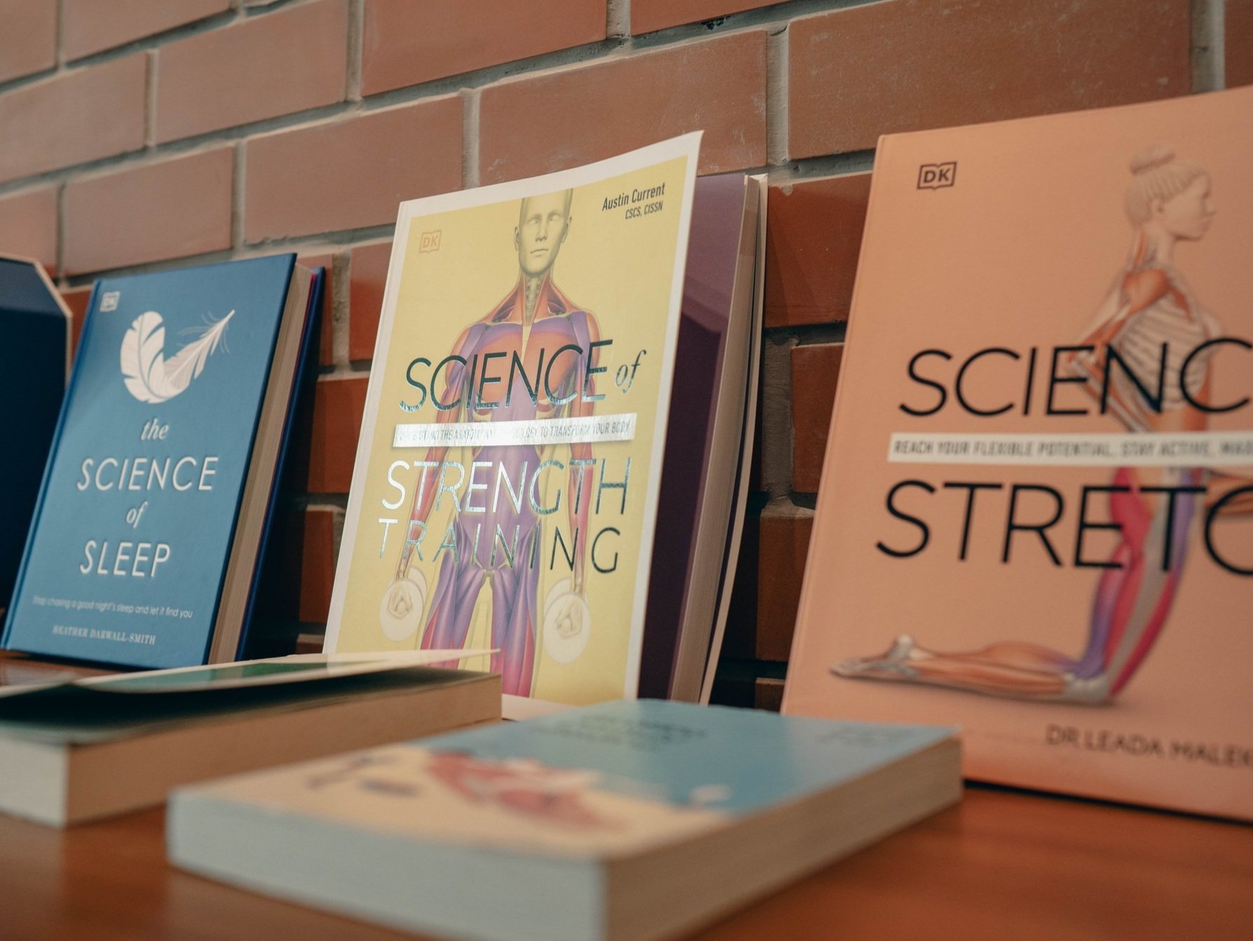Three books on a wooden table in front of a brick wall, with two more books laying flat. The books are about science topics, including sleep, strength training, and stretching.