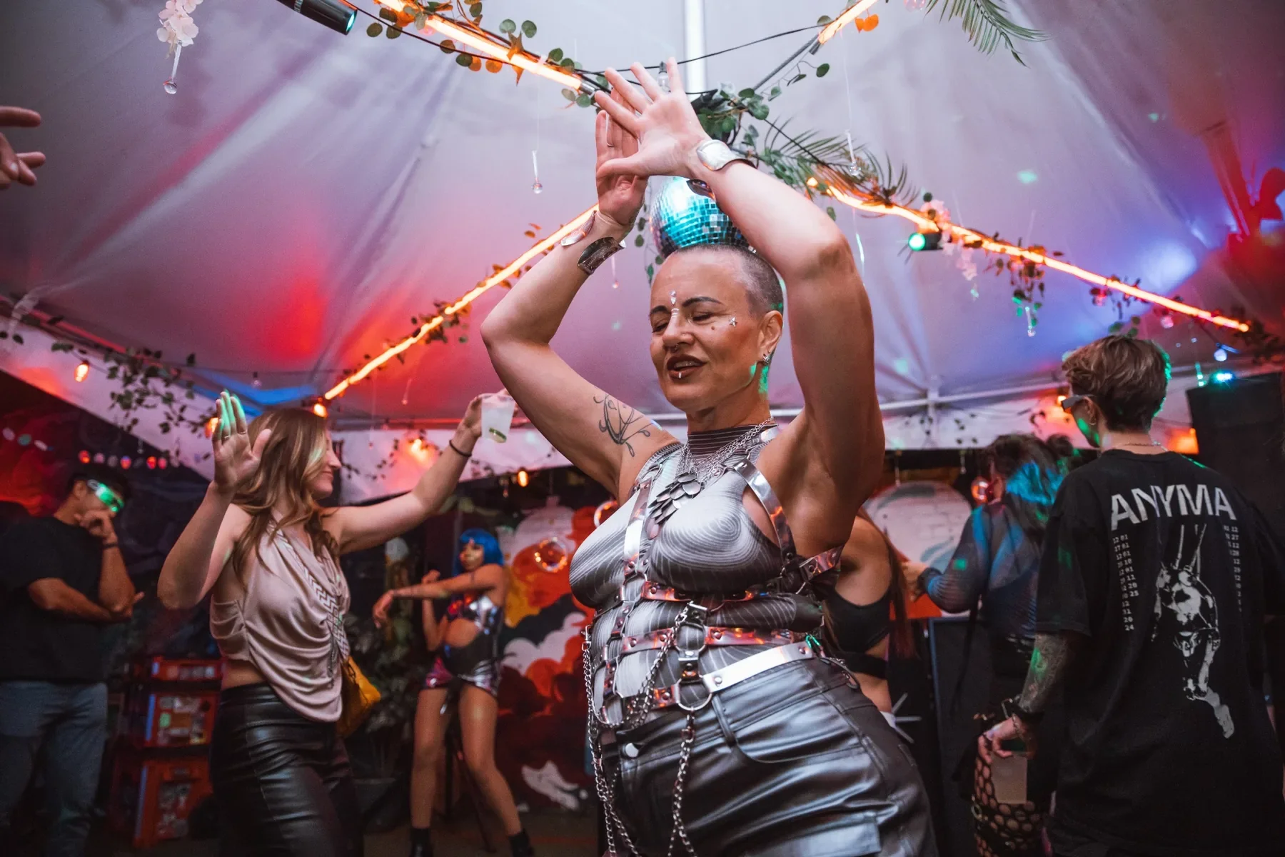 Guest in a silver outfit dances with raised arms on the Cyber Noir dance floor beneath a disco ball.