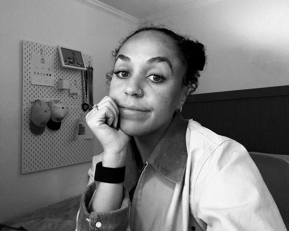 A young woman with curly hair in two buns, wearing a nose ring and earrings, sitting in a room. She rests her chin on her hand, looking into the camera. There is a pegboard with hanging hats, pictures and metals in the background.