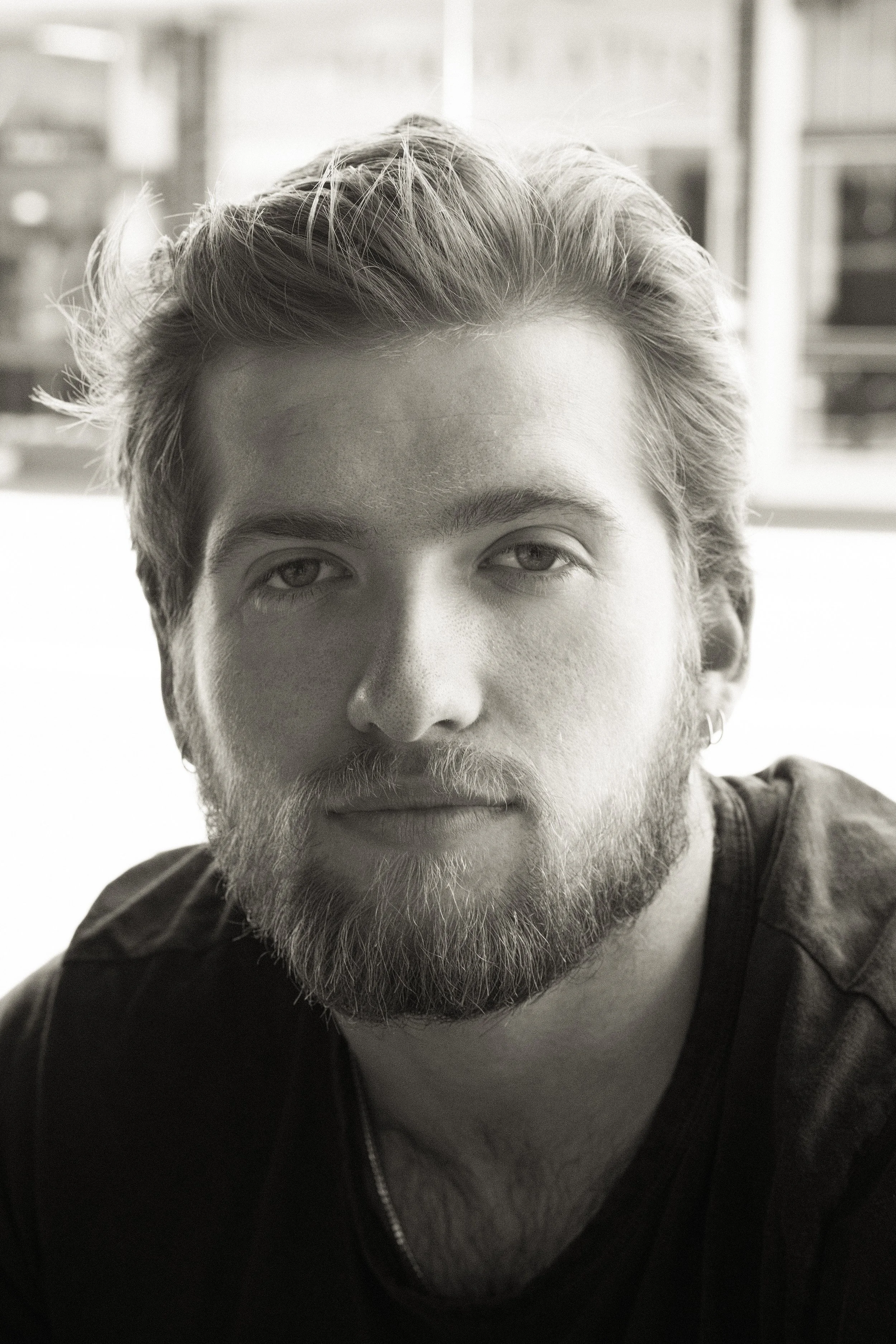 A young man with tattoos and jewelry, wearing a tank top, resting his elbows on his knees, looking directly at the camera against a plain background.