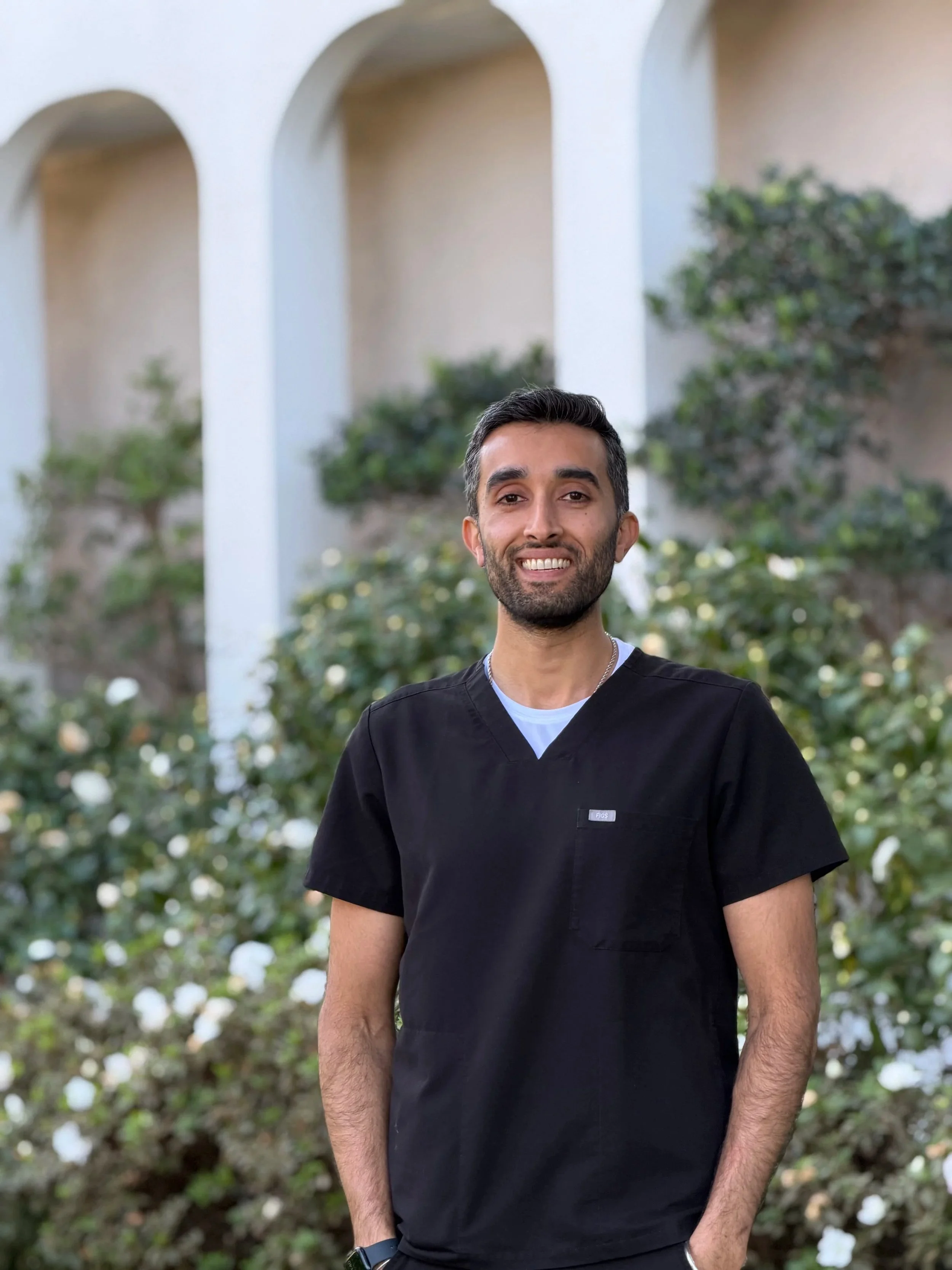 Professional picture of Dr. Arvin Pal, orthodontist, wearing black Figs scrubs and standing in front of a row of white arches with greenery and white flowers.