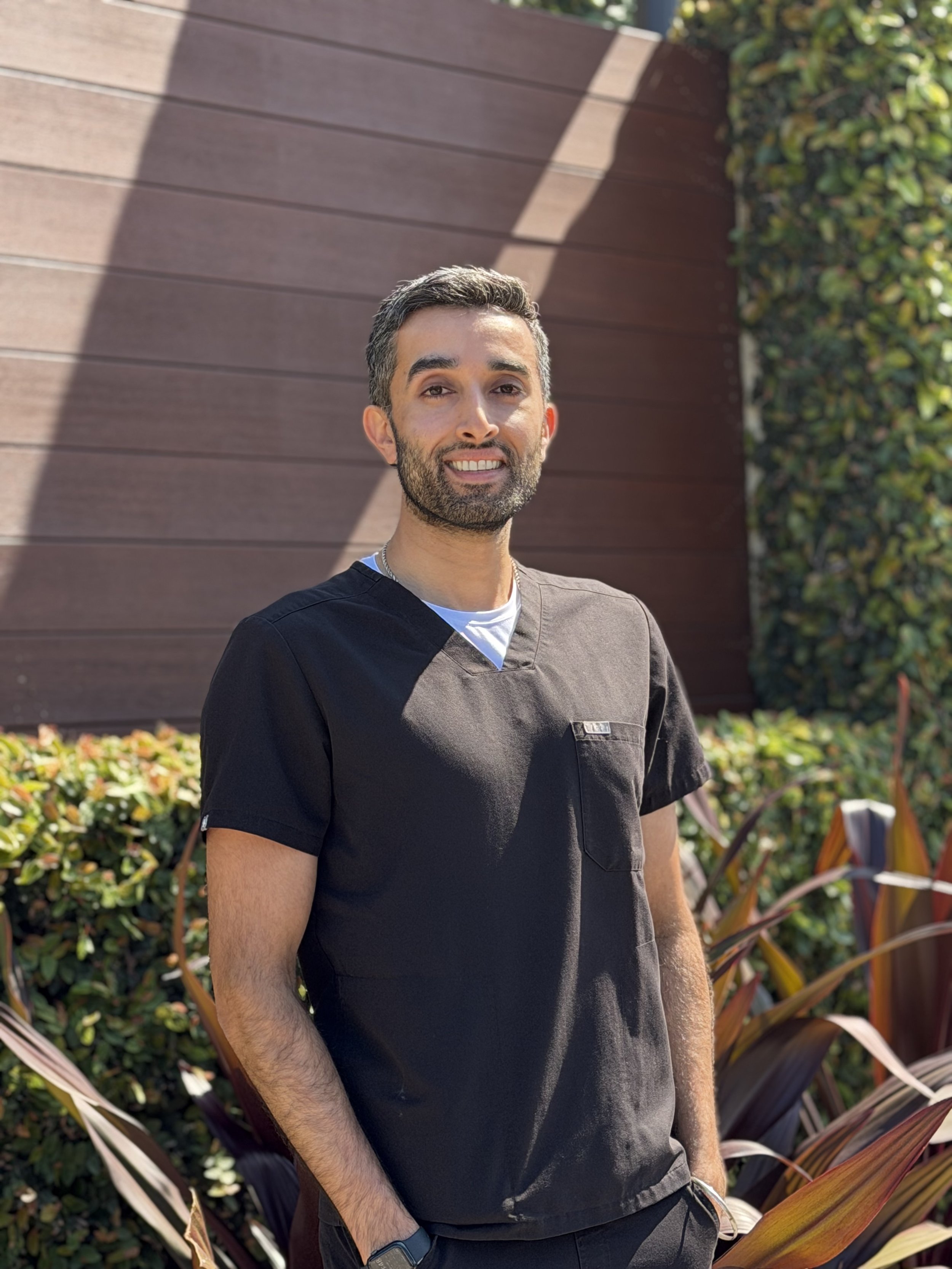 Professional picture of Dr. Arvin Pal, orthodontist, wearing black Figs scrubs and standing in front of a wooden wall with greenery and bushes.