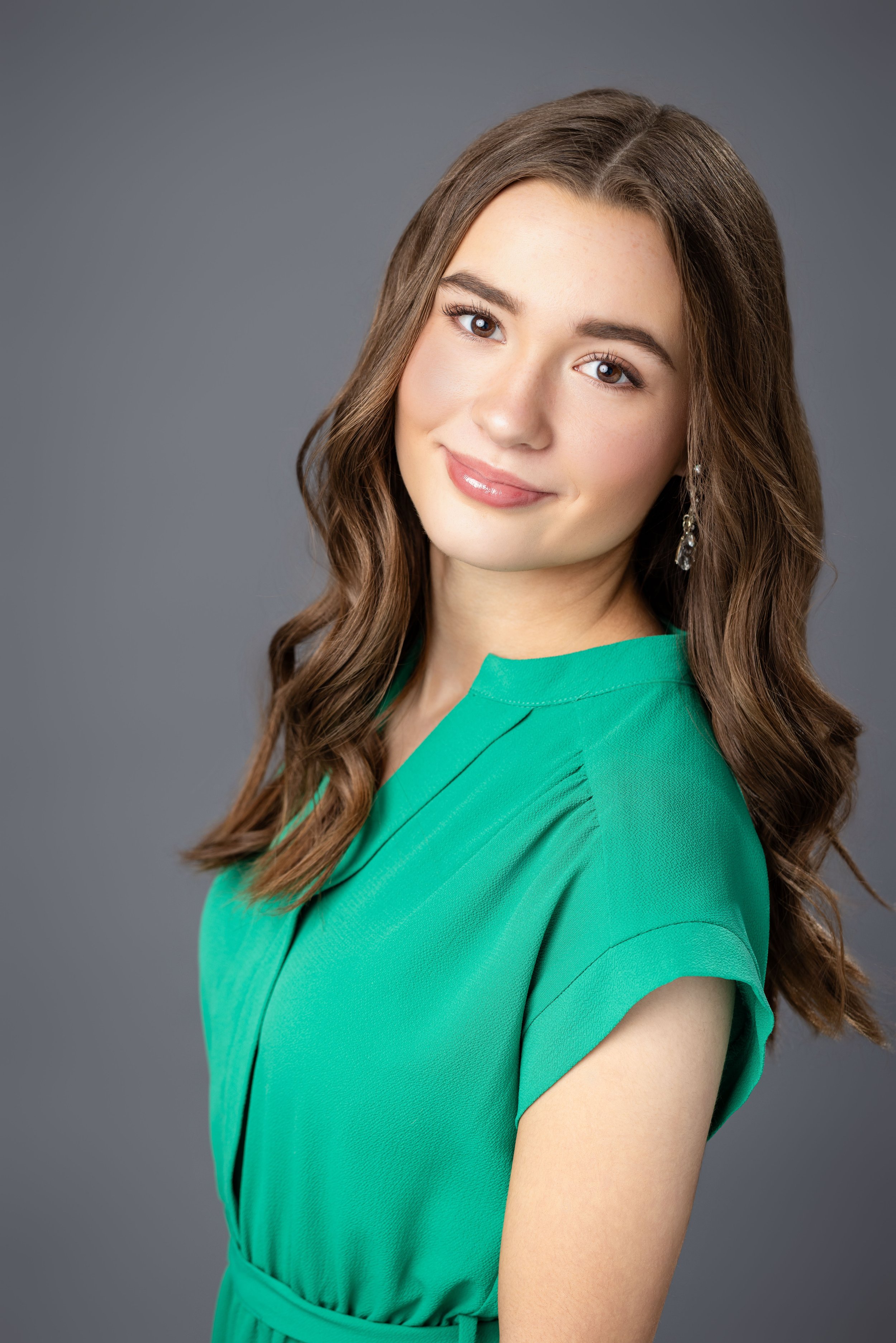 Head-and-shoulders portrait of a young woman with long brown hair wearing a green top and earrings, smiling softly against a gray background.