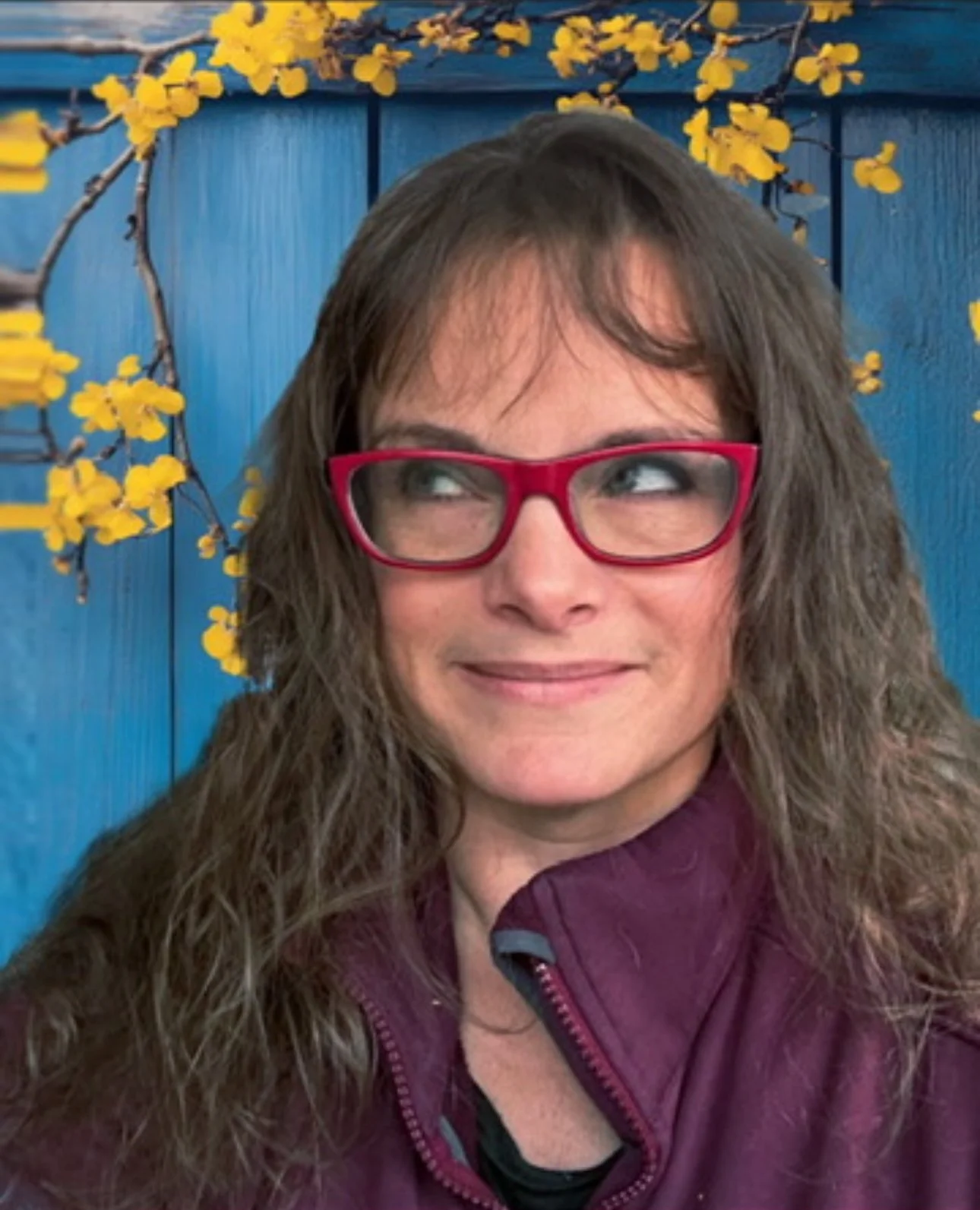 A woman with long, wavy brown hair wearing pink glasses and a maroon jacket standing in front of a blue fence decorated with yellow flowers.