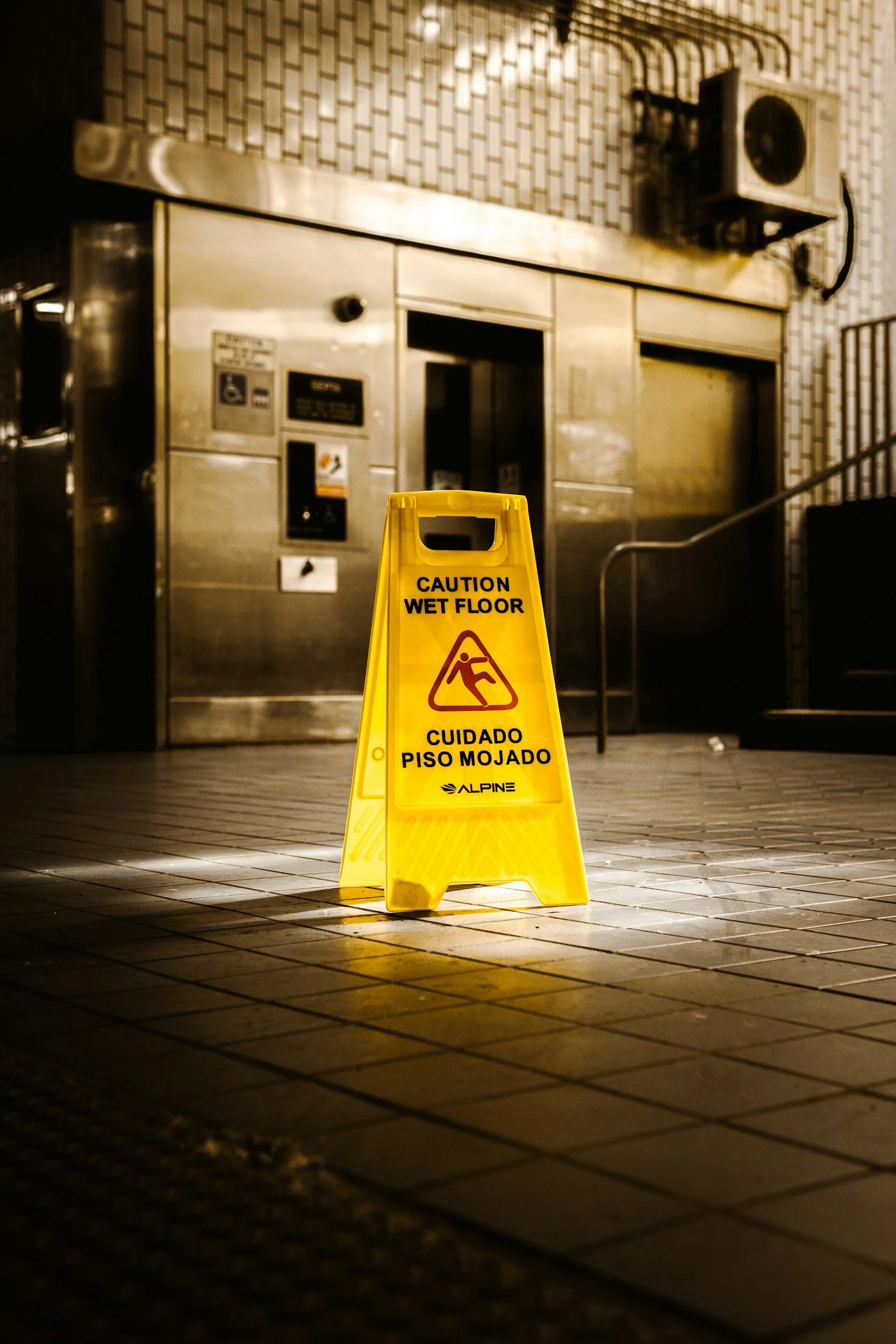 A yellow caution wet floor sign in the foreground on a tiled floor. In the background, there is an elevator with metallic doors and a tiled wall, and an air conditioning unit mounted near the ceiling.