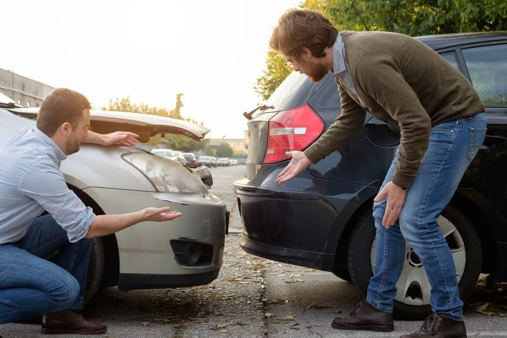 Two men are arguing over a minor car accident in a parking lot, with one man pointing towards a damaged white car and the other towards a black car.