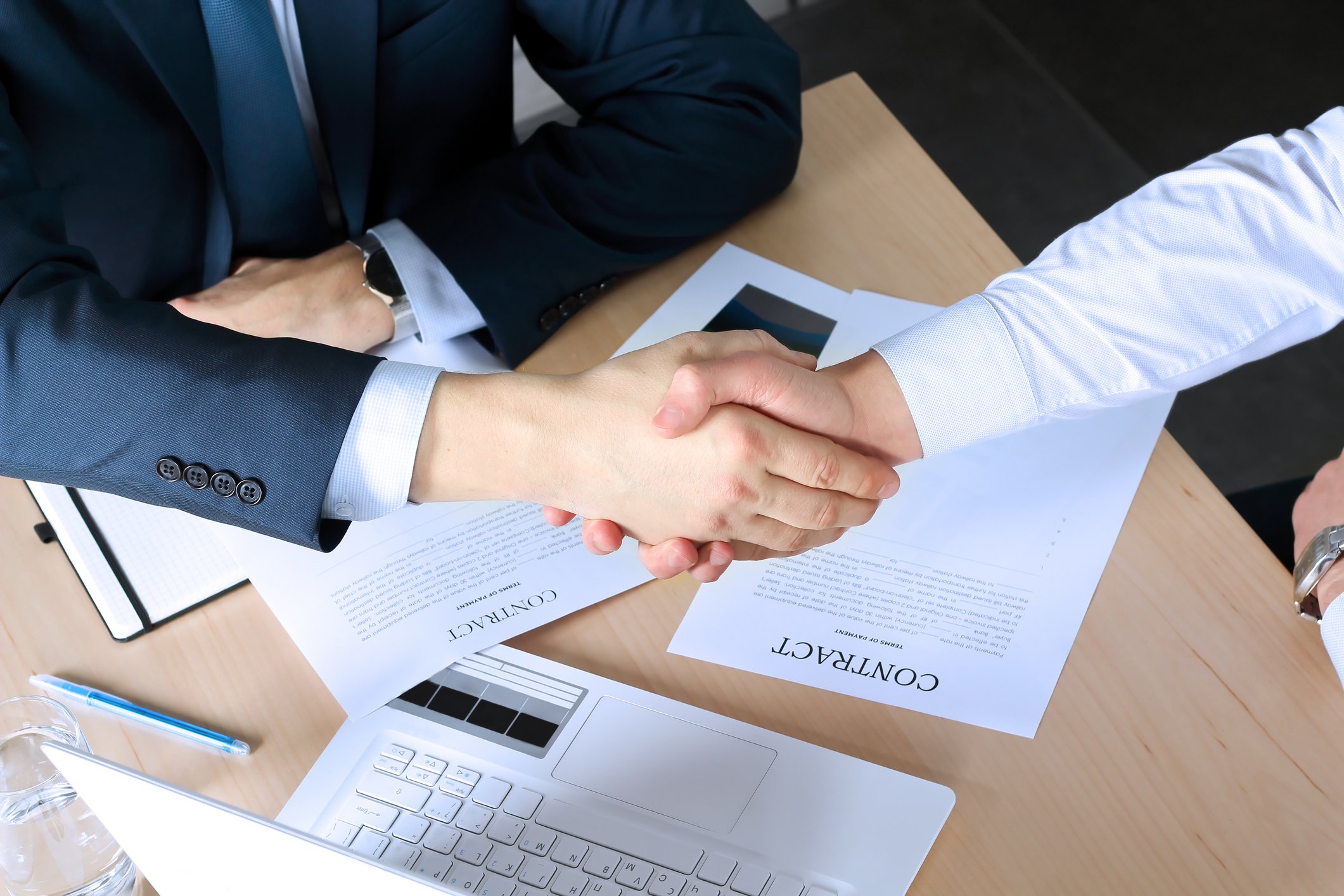Two people shaking hands over a meeting table with a contract, documents, pen, laptop, and glass of water on the table.