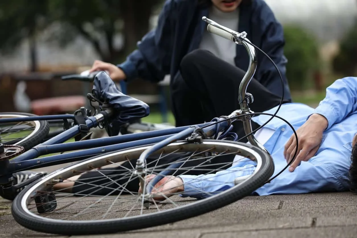 Person lying on the ground with bicycle, another person kneeling nearby, outdoors.