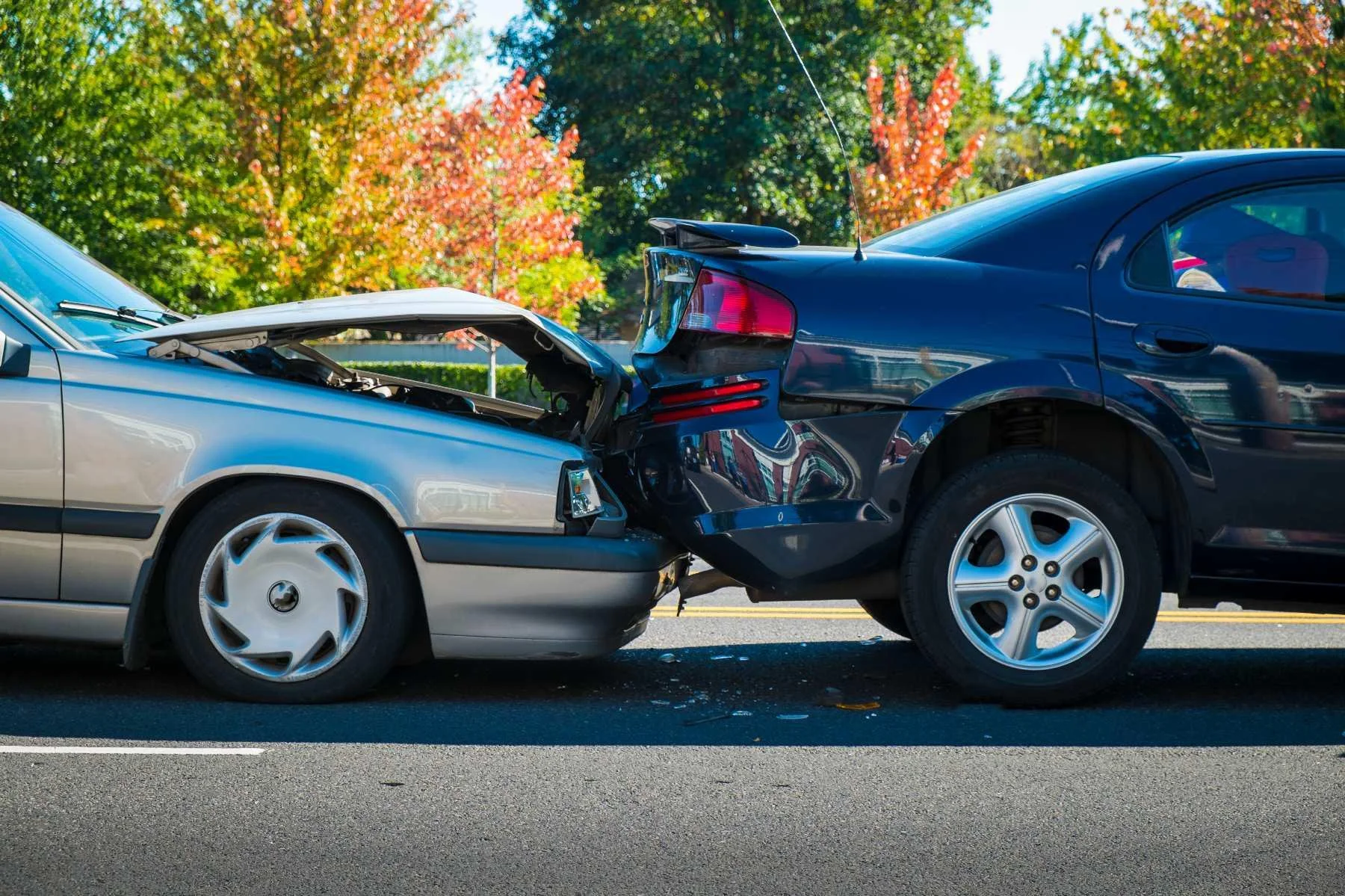 Two cars involved in a collision on the road. The front of a silver sedan has collided with the back of a dark blue vehicle, with visible damage and debris on the road. Trees with autumn foliage are in the background.