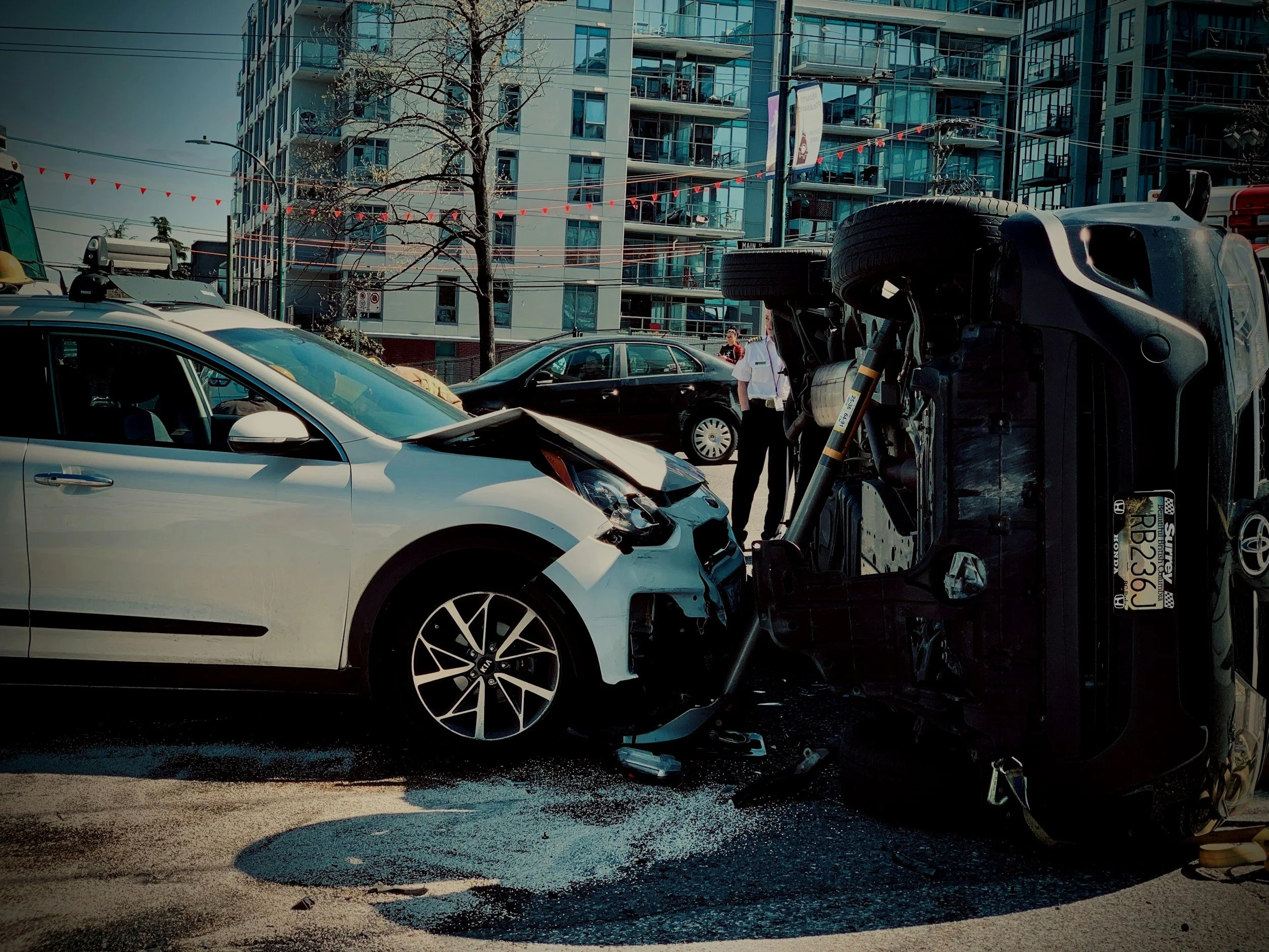 A white car involved in a car accident with a black vehicle lying on its side, and a damaged front end with a shattered windshield. In the background are modern apartment buildings, a tree, and people on the street.