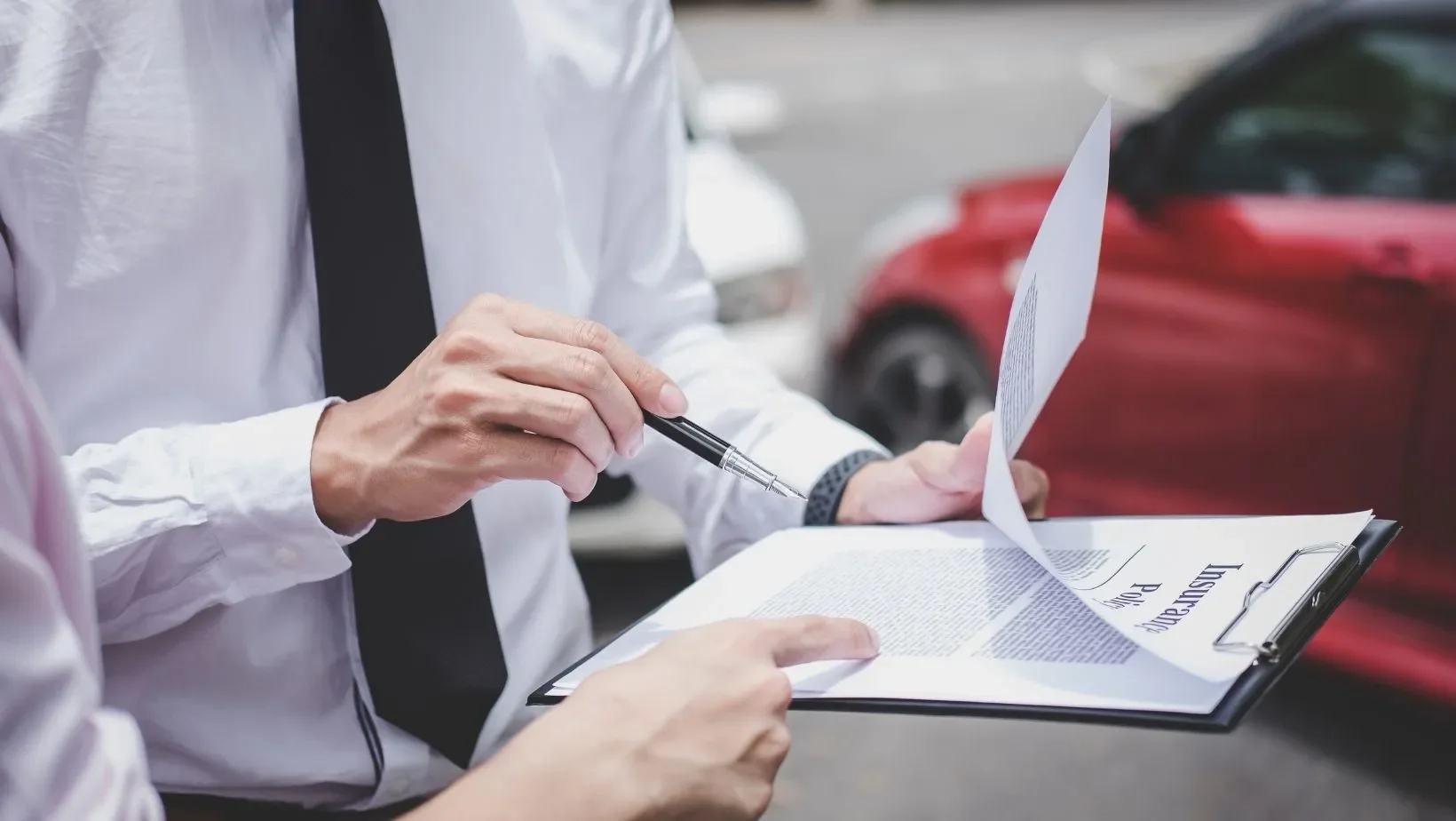 Close-up of two individuals, one pointing at a document with a pen, while the other holds a clipboard with a paper on it, outdoors near parked cars.