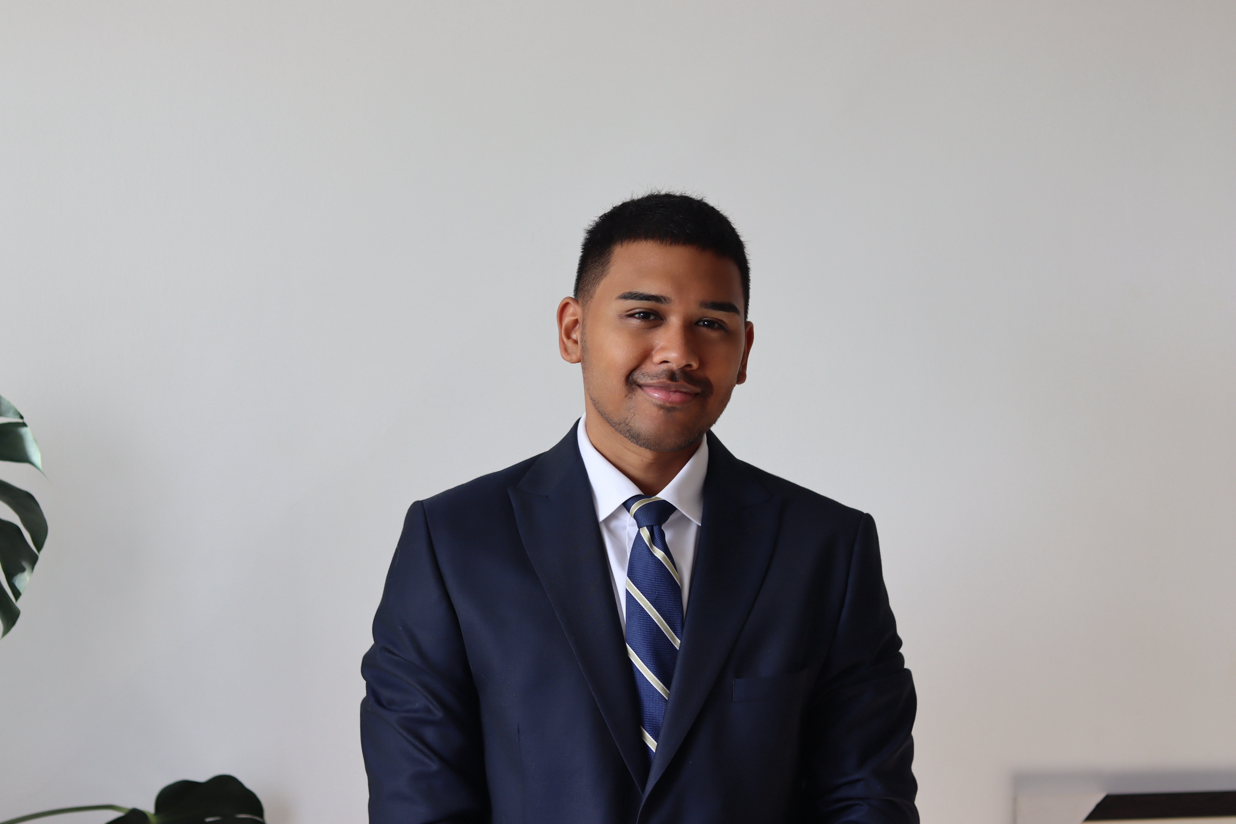 Portrait of a young man wearing a dark suit, white shirt, and striped tie, smiling at the camera against a plain light-colored background with a green plant partially visible on the left.