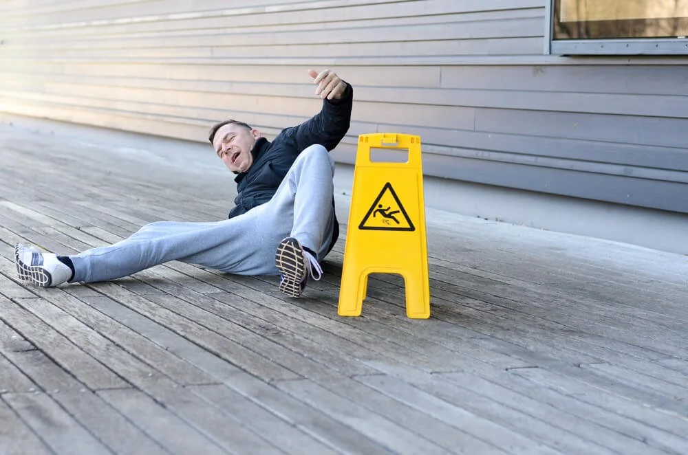Man on the ground smiling near a "wet floor" warning sign outside a building.