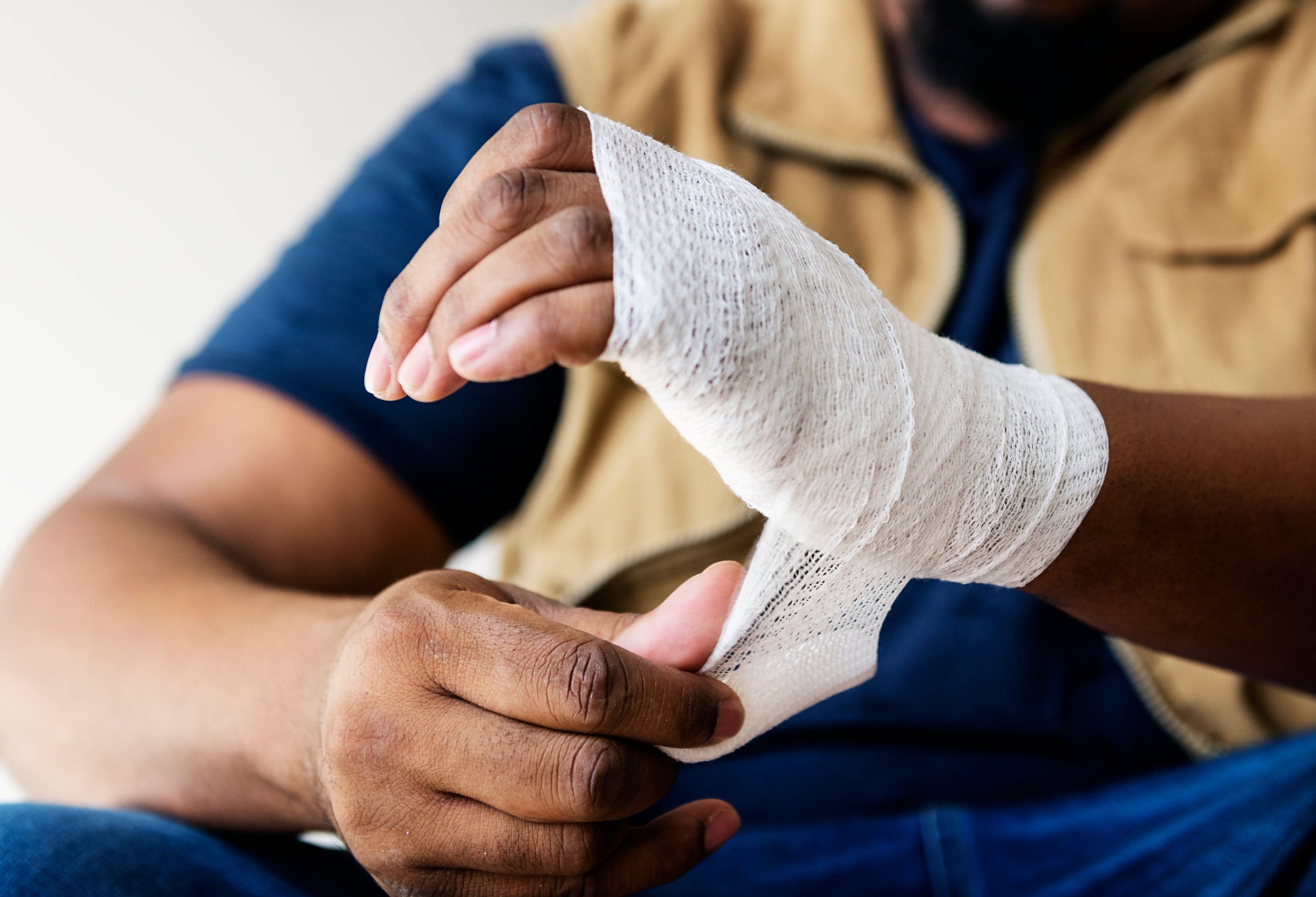 A person with dark skin is holding a white bandaged hand, with the background showing parts of their blue shirt and beige jacket.