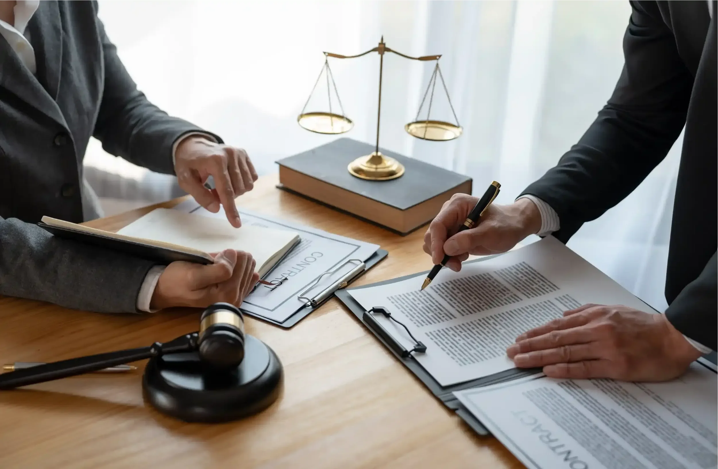 Two individuals in business attire reviewing legal documents at a wooden table, with a gold-scale balance model and a telescope in the background.