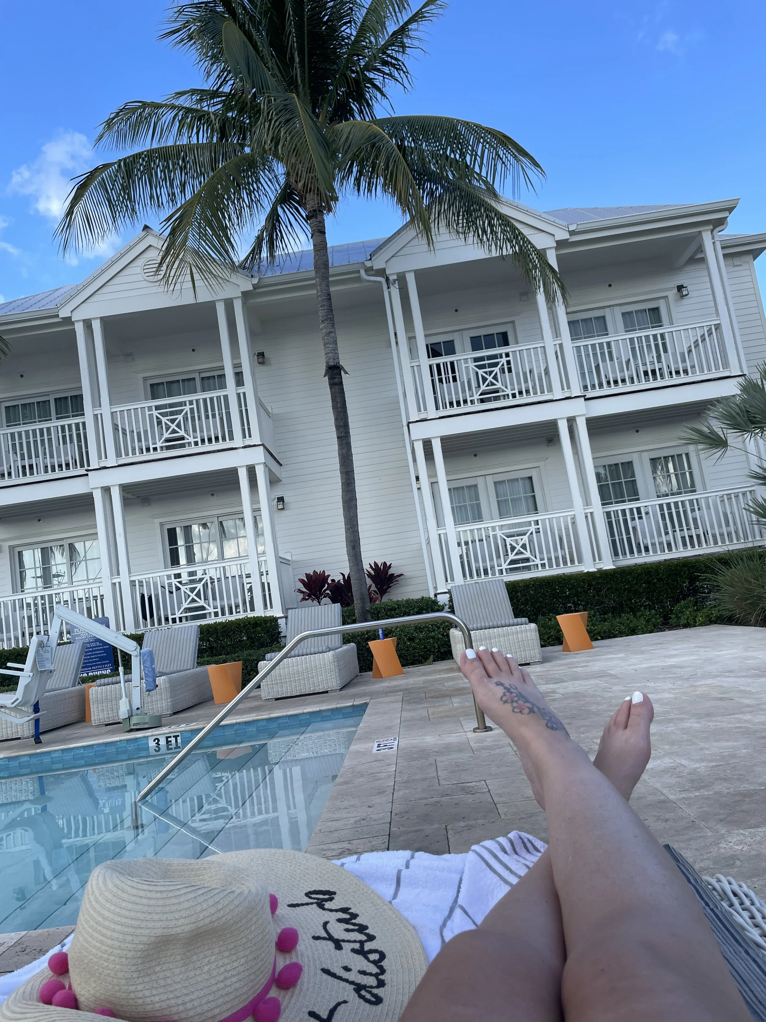 View of a poolside area at a hotel or resort with a white multi-story building in the background, balconies, lounge chairs, a woman with tattooed legs relaxing by the pool, a sunhat with pink pom-poms, and a palm tree, under a clear blue sky.