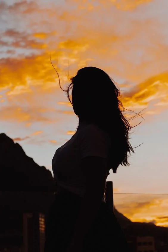 Silhouette of a woman standing outdoors during sunset with a scenic sky and mountains in the background.