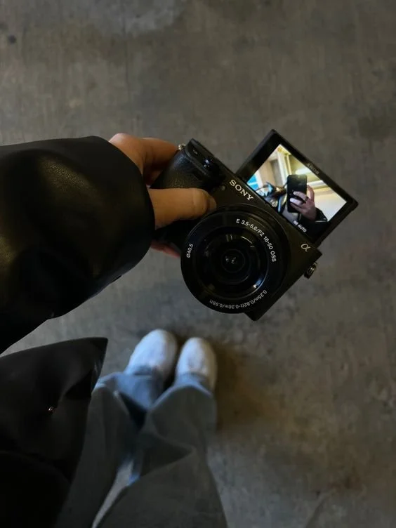 Person taking a photo with a Sony digital camera, capturing their reflection on the camera's screen. The person is wearing light-colored shoes and dark clothing, standing on a textured gray floor.