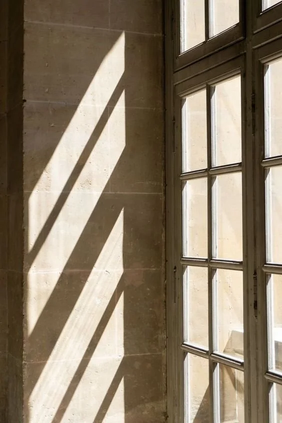 Sunlight casting diagonal shadows through window panes onto a beige stone wall.