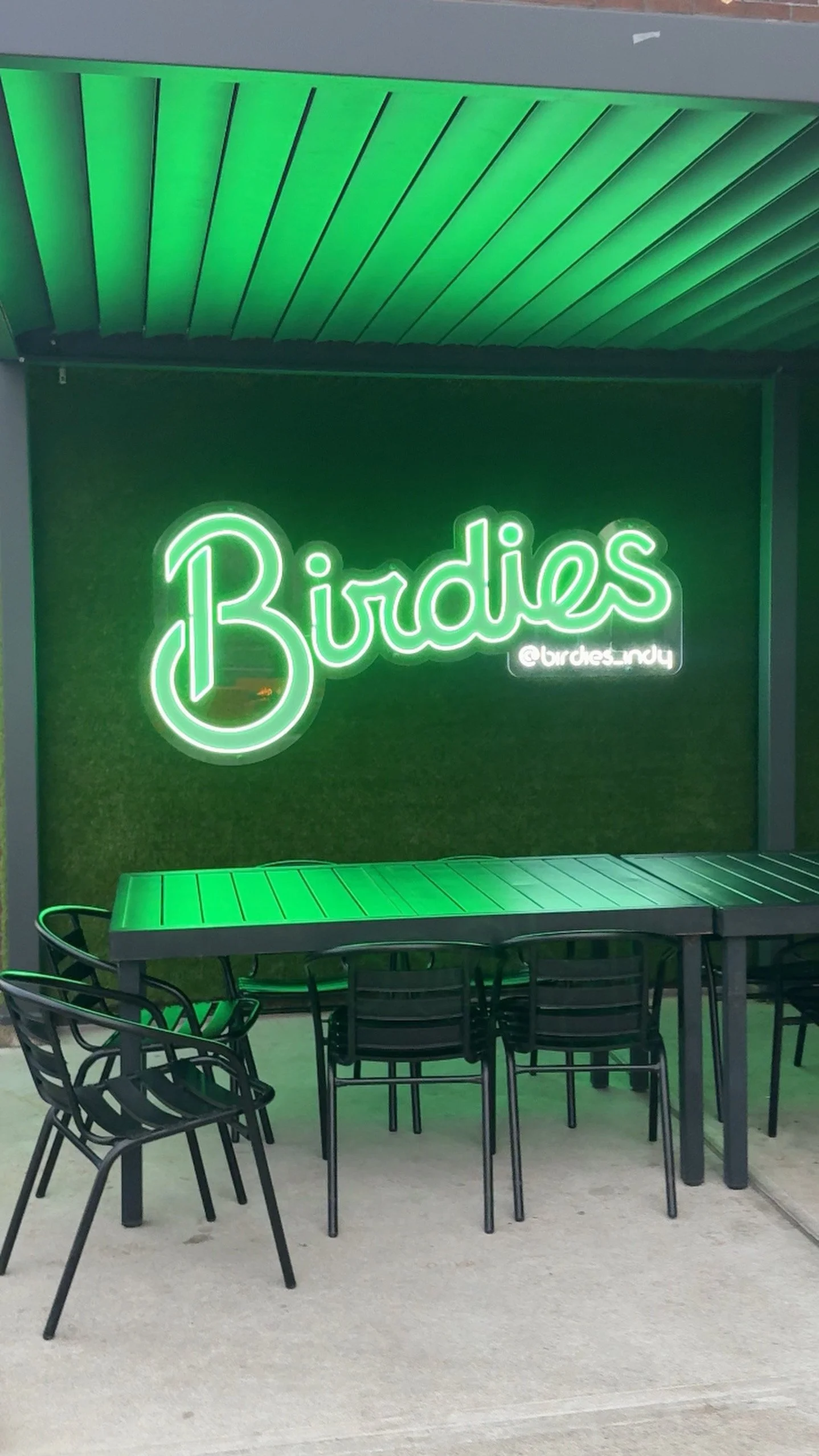 A neon sign with the word 'Birdies' in green cursive font and the Instagram handle '@birdies.indy' underneath. The sign is mounted on a green wall with a small table and black chairs in front.
