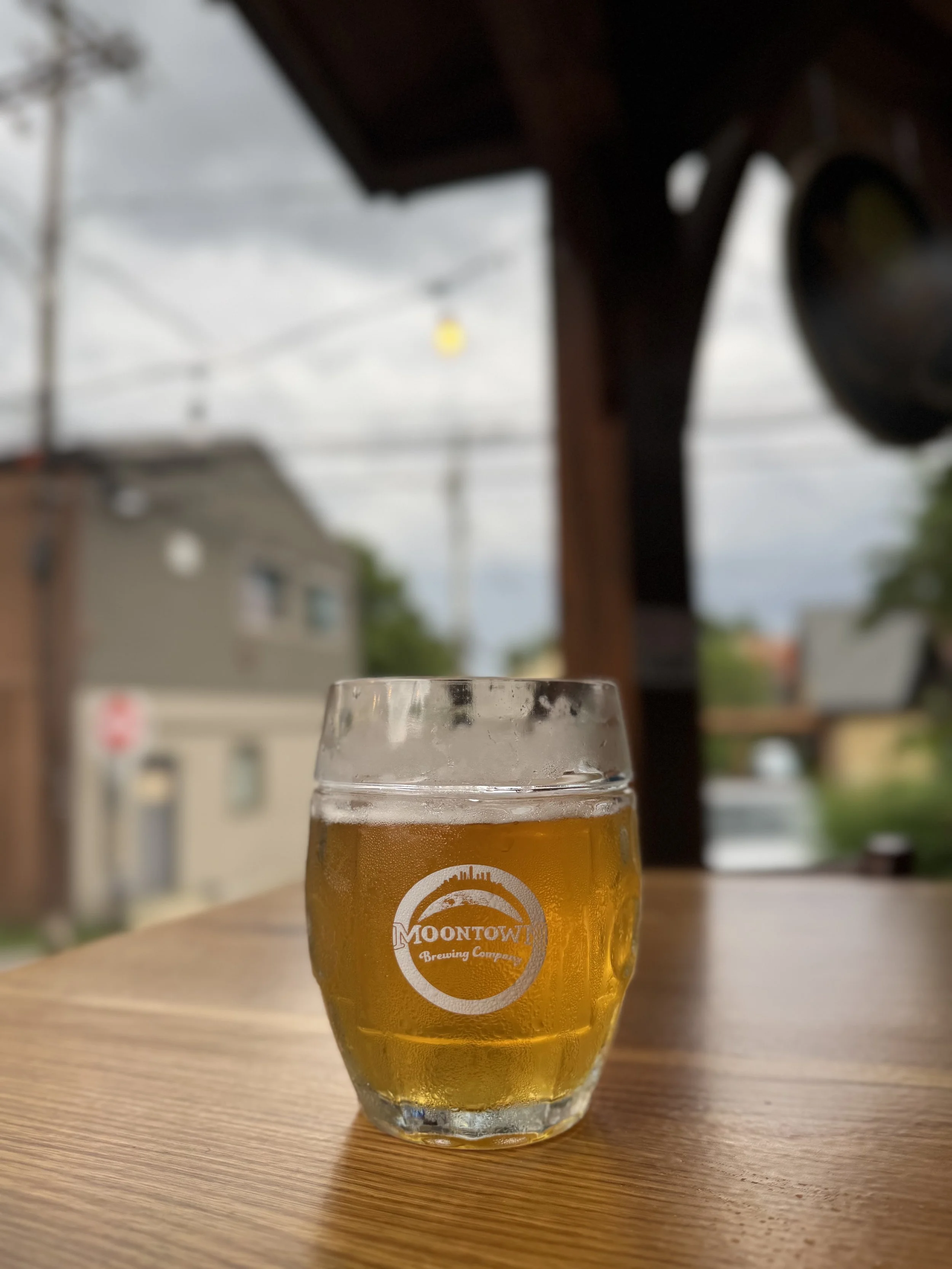 A glass of beer with a logo that reads 'Montow Brewing Company' on a wooden table, with a blurred outdoor background showing trees, buildings, and power lines.