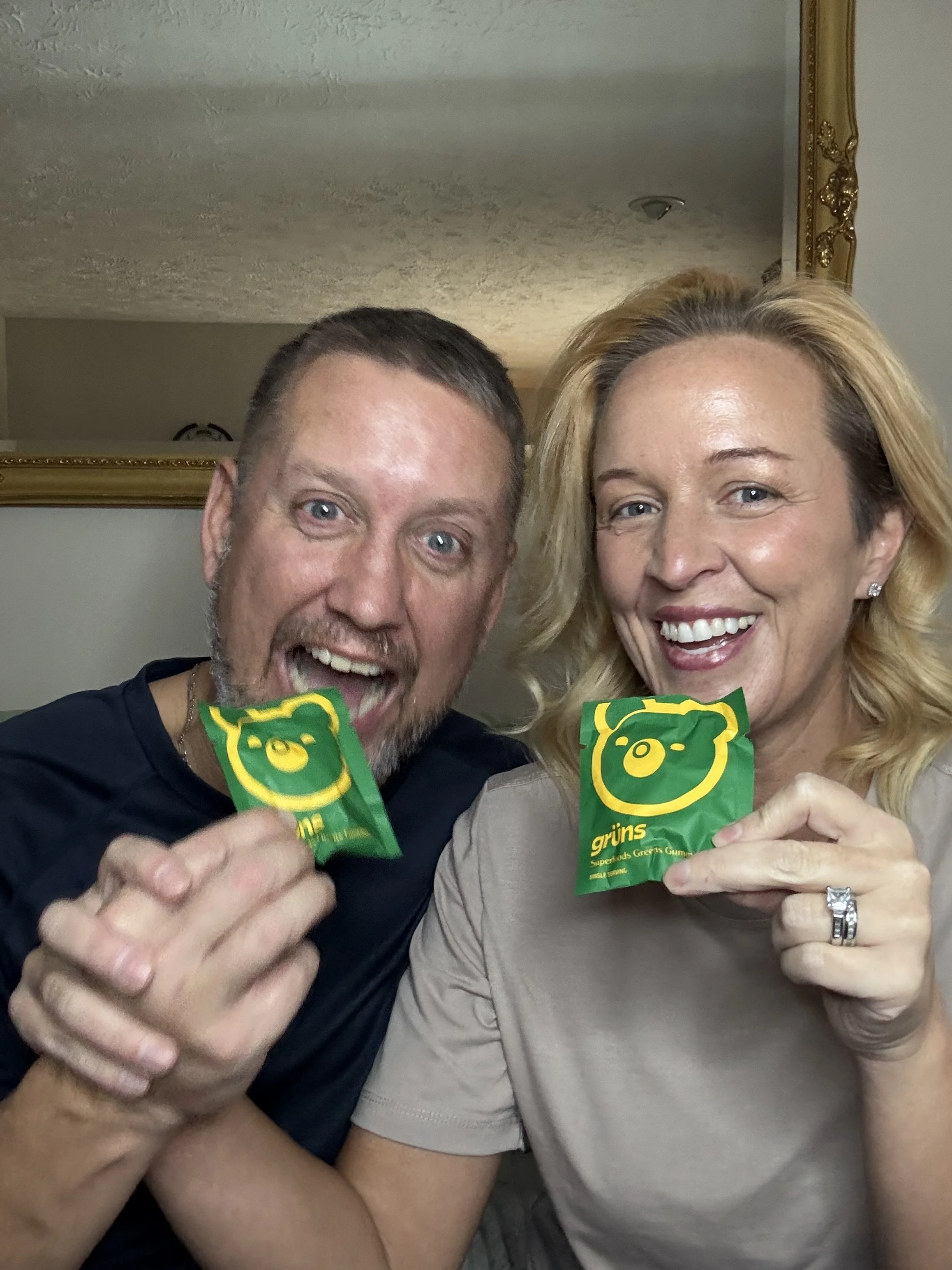 Two smiling people, a man and a woman, are holding green candy bags with a yellow bear face and the word 'grüns' on them. They appear to be happy and are in a close-up selfie.