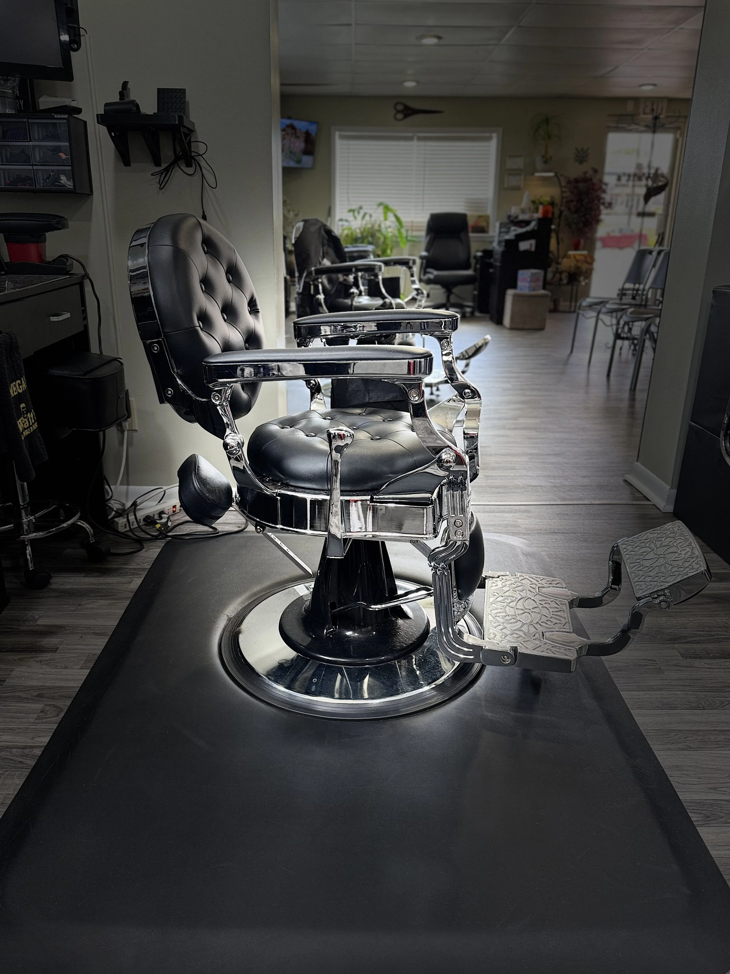 Barber's chair in a barbershop with a black leather and chrome design, positioned on a black countertop, with various hair and grooming tools in the background.